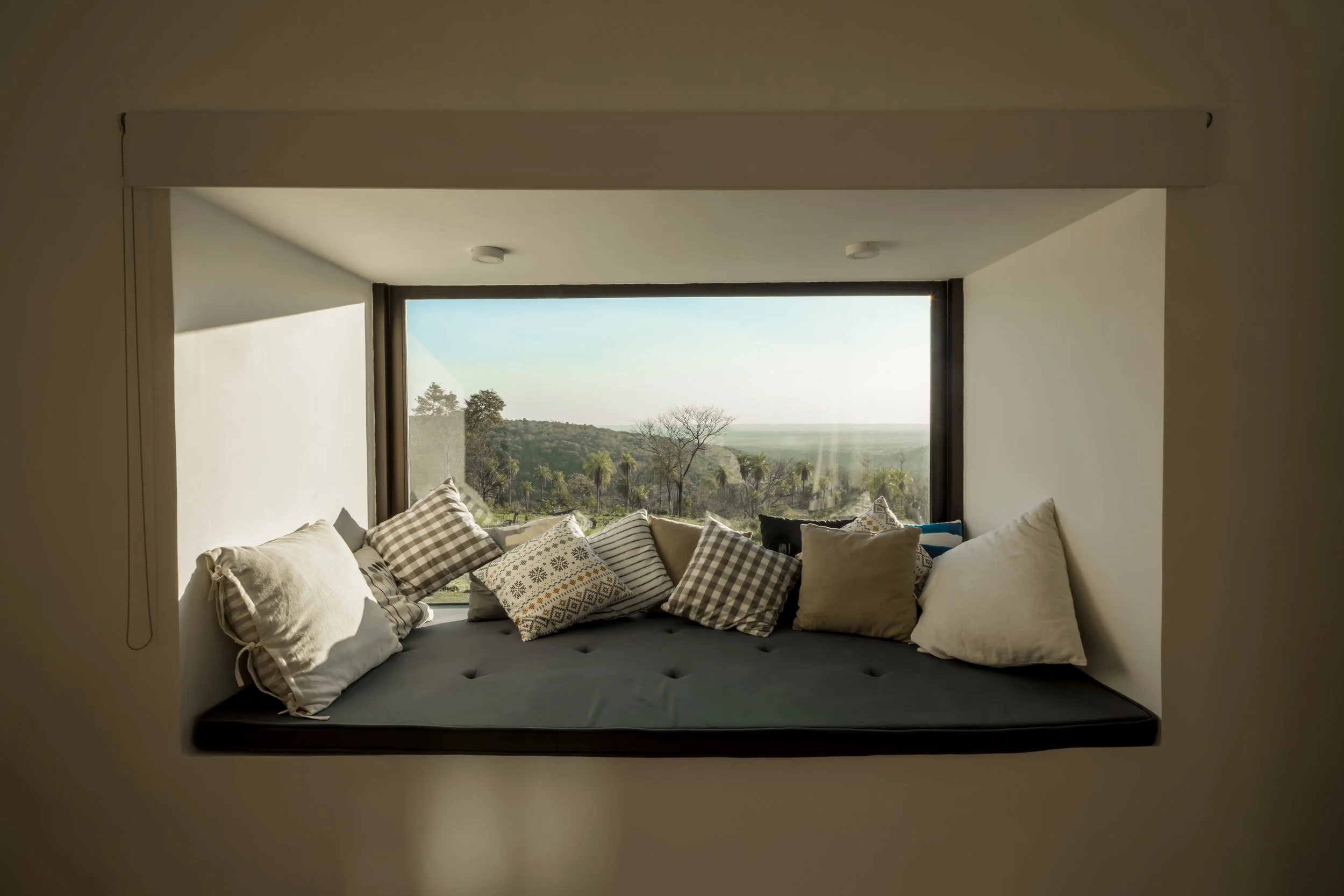 PRO ARQUITECTURA PARAGUAY architecture design PROSA
Ventana grande con almohadones de diferentes colores y patrones en un asiento junto a la vista de un paisaje natural con árboles y colinas.