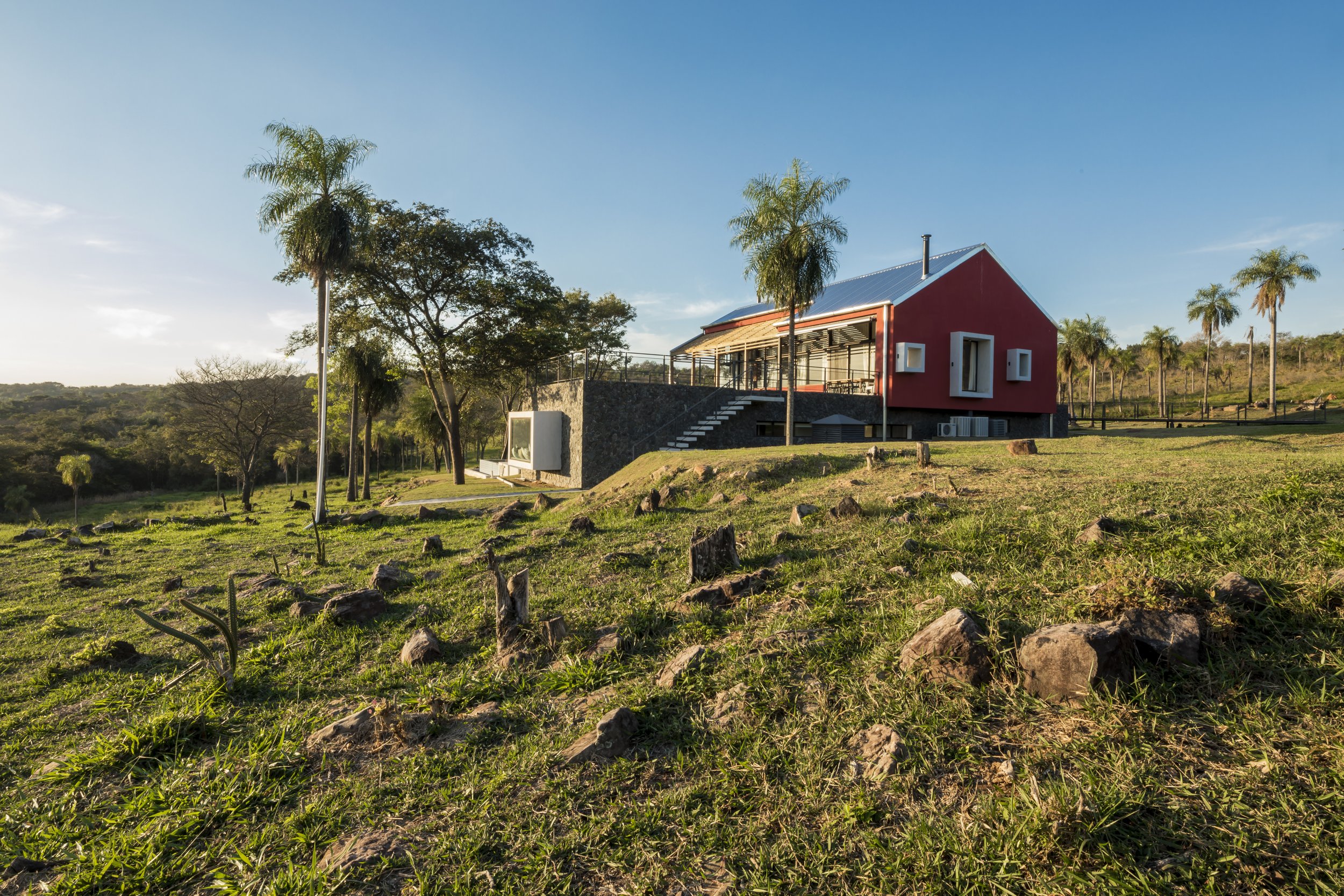 PRO ARQUITECTURA PARAGUAY architecture design PROSA
Casa moderna de color rojo con terraza en una colina, rodeada de árboles y palmeras, bajo un cielo despejado