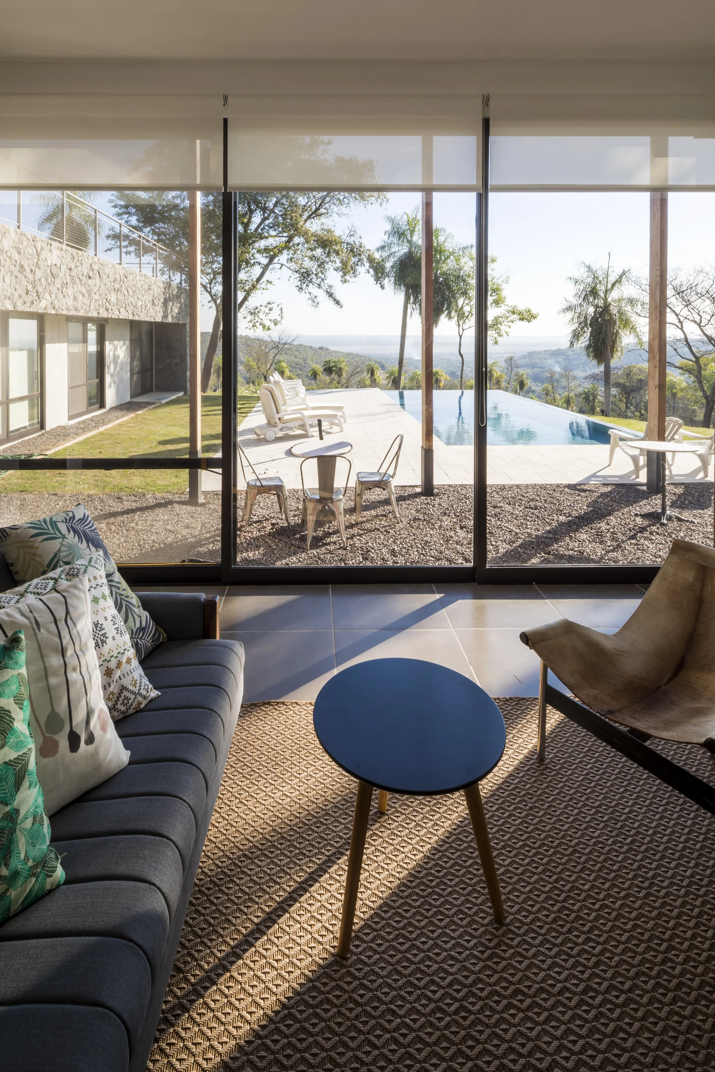 PRO ARQUITECTURA PARAGUAY architecture design PROSA
Vista de una sala interior con ventanas grandes que muestran una piscina y un paisaje con árboles y colinas.