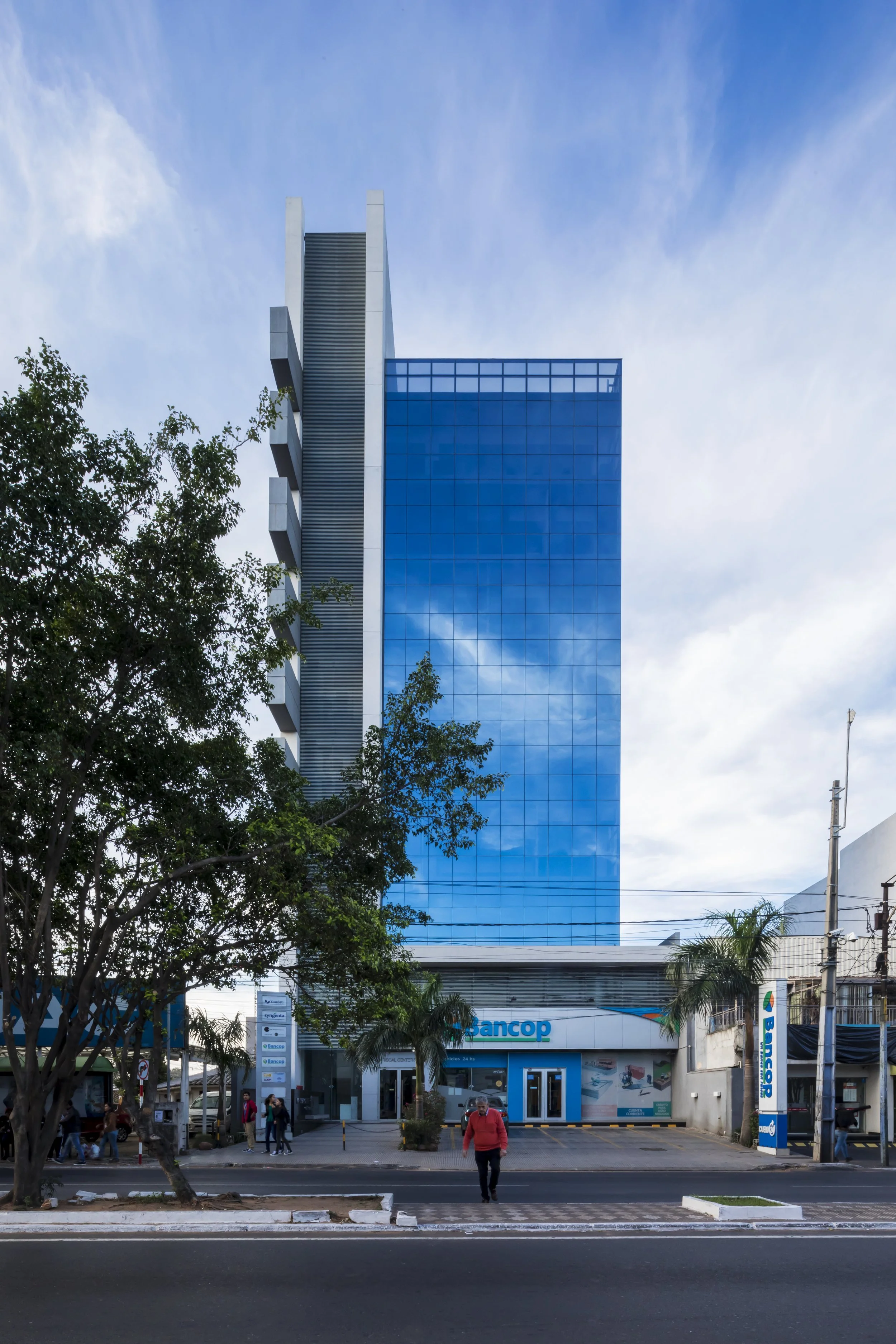 PRO ARQUITECTURA PARAGUAY architecture design PROSA
Edificio alto de vidrio azul con un letrero de Bancop en la entrada, árboles y personas caminando frente a la estructura bajo un cielo parcialmente nublado.