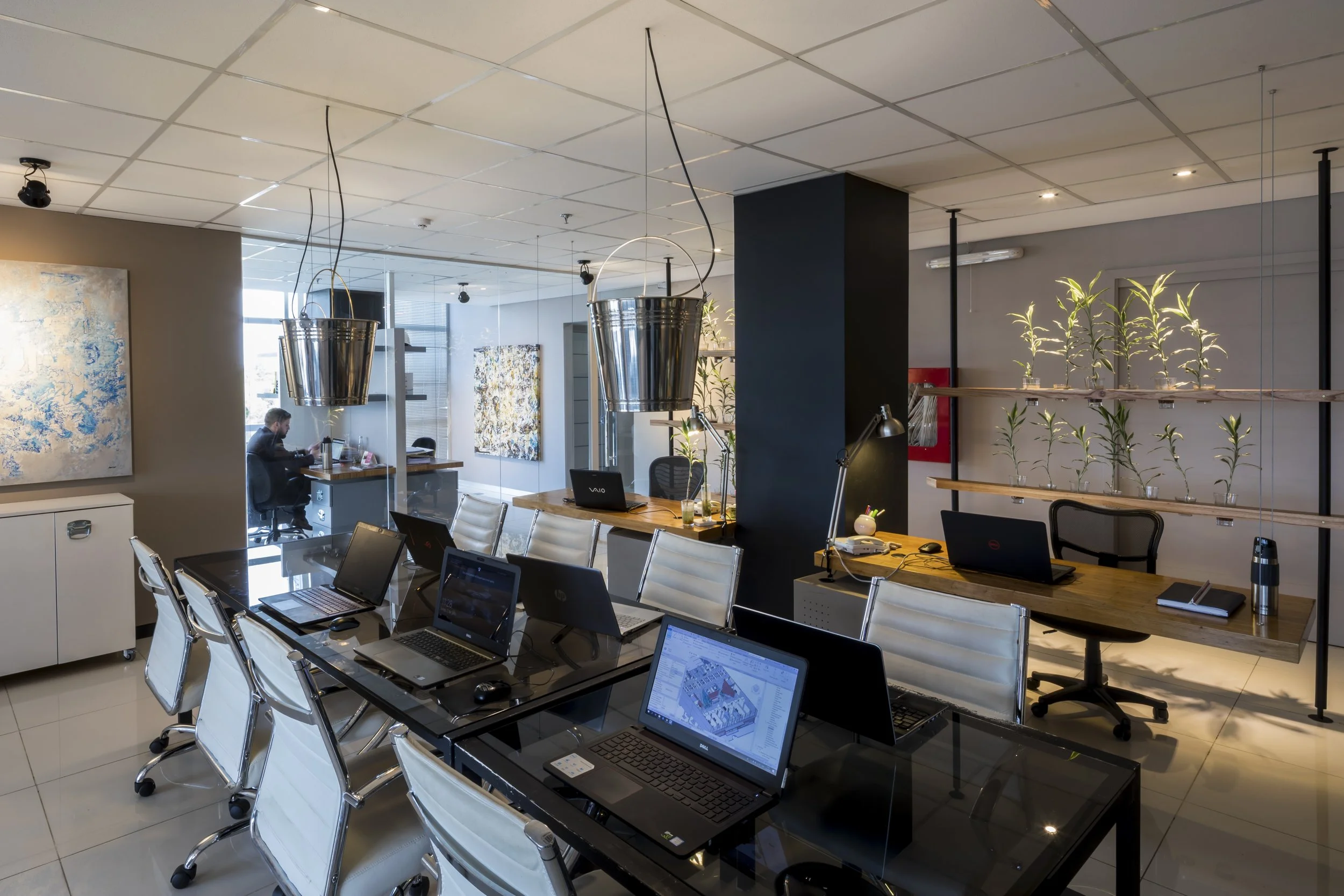 Sala de conferencias moderna con varias computadoras portátiles en una mesa grande y sillas blancas. En el fondo, hay una pared con plantas enmarcadas y una persona trabajando en un escritorio. Decoración con pinturas abstractas en las paredes y lámp