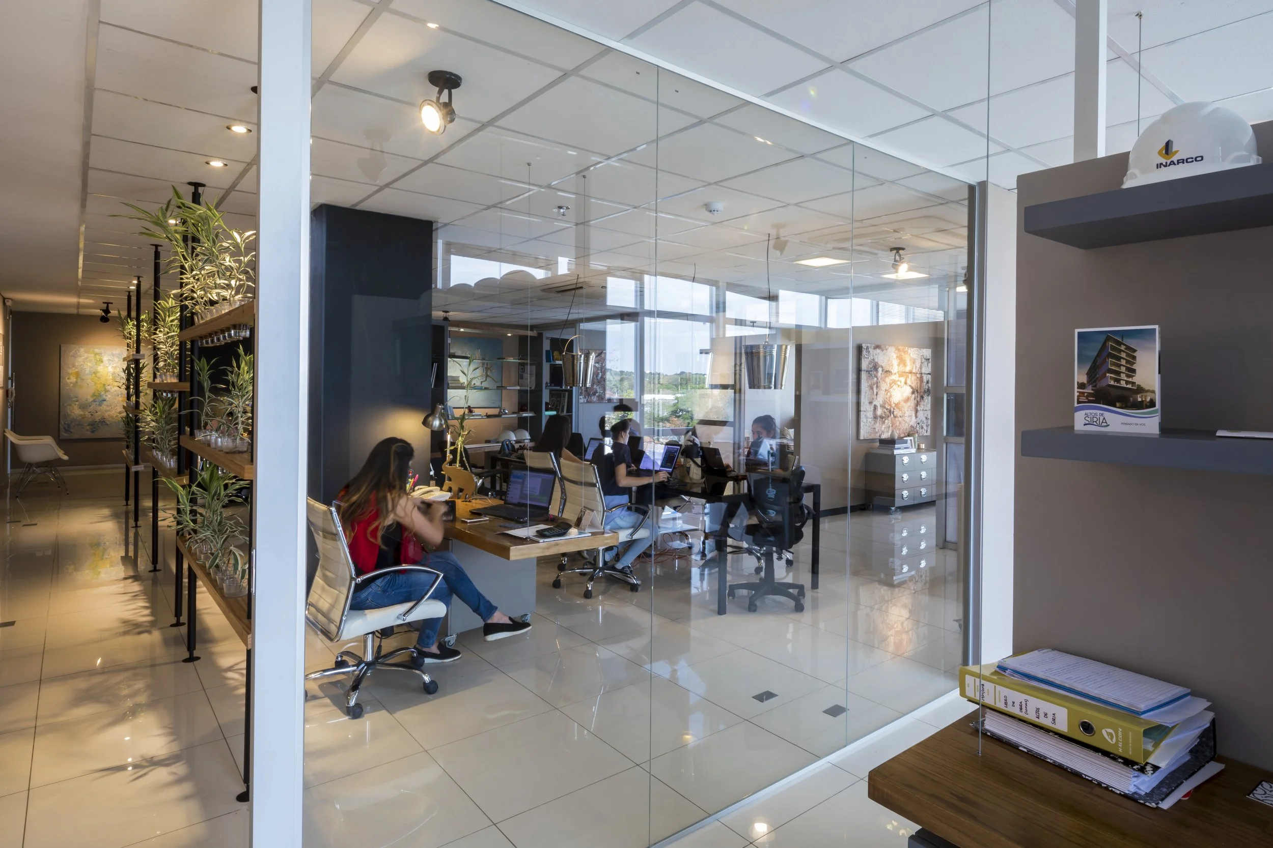 Vista interior de una oficina moderna con varias personas trabajando en sus computadoras, vista a través de un muro de cristal. Hay plantas decorativas y piezas de arte en las paredes.