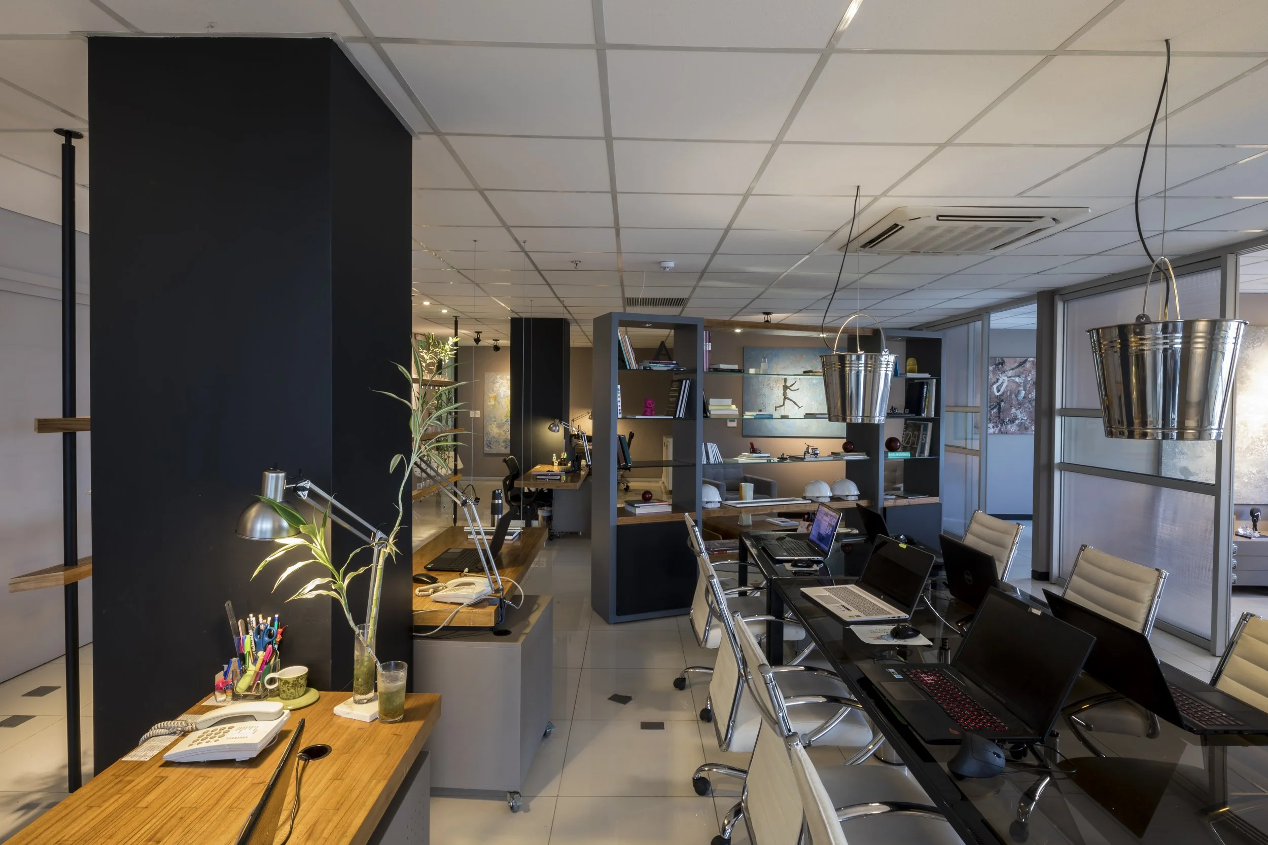 Oficina moderna con varias mesas de trabajo, sillas blancas, computadoras portátiles, estanterías con libros y accesorios decorativos, ventanas grandes con vista a árboles y cuadros en las paredes.