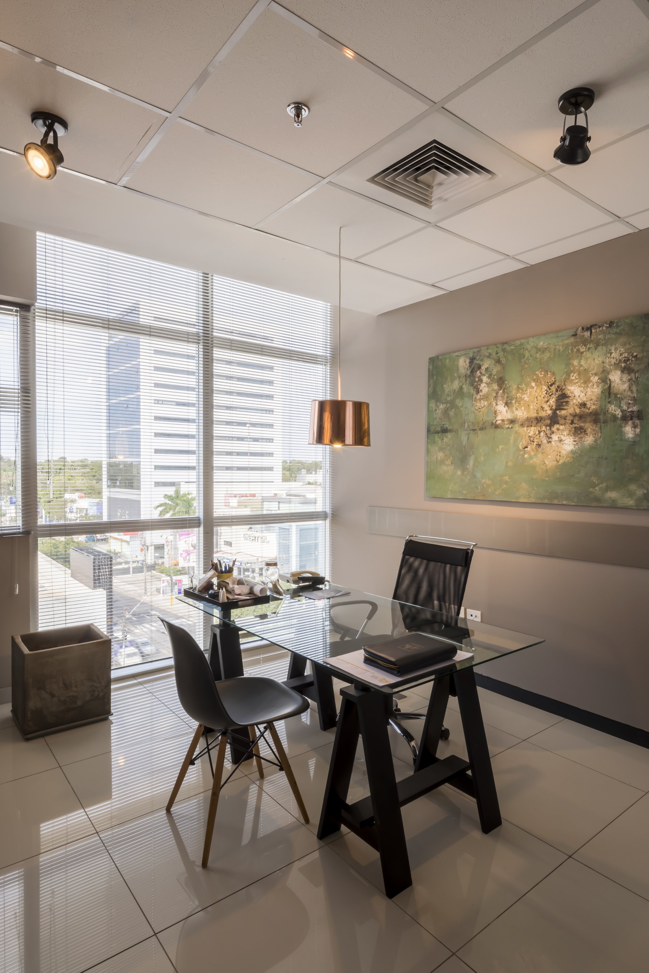 Oficina moderna con mesa de vidrio, sillas, cuadro abstracto en la pared y ventanas grandes con vista a edificios de la ciudad.