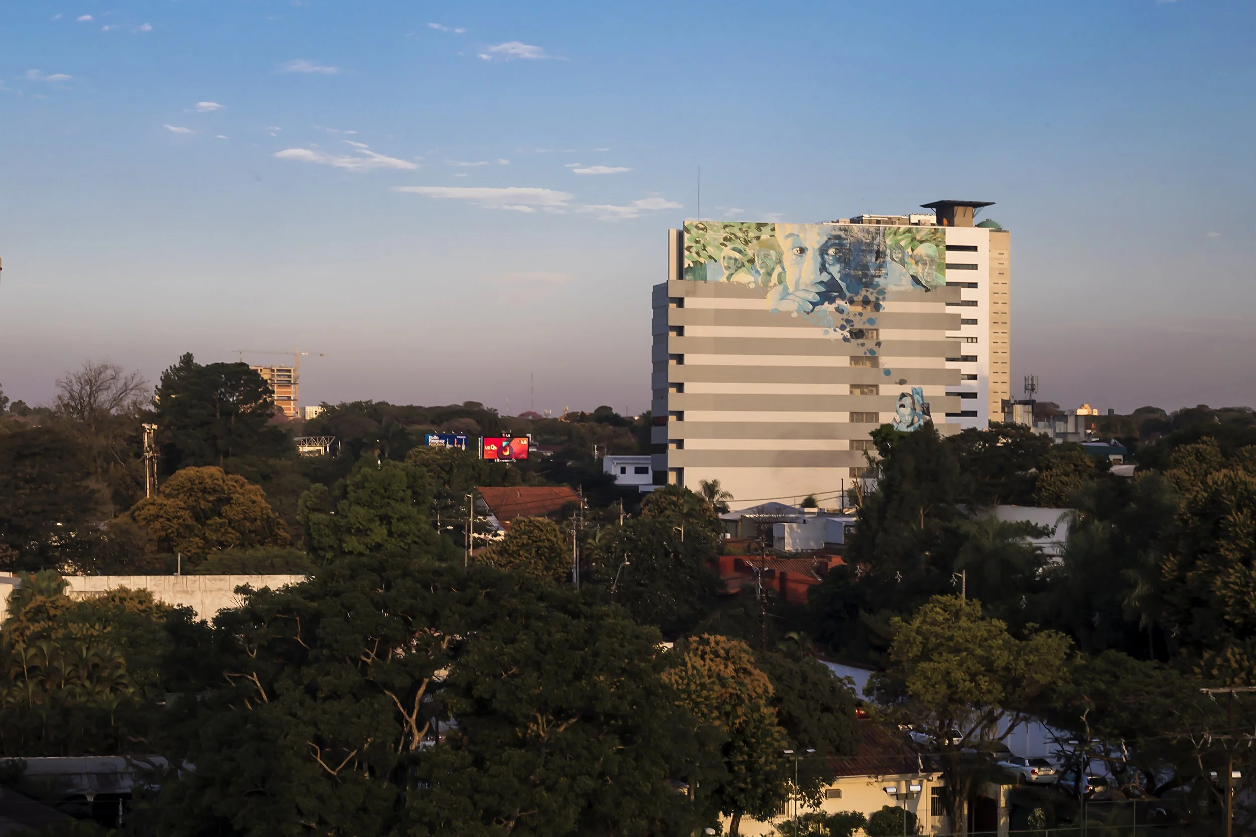 PRO ARQUITECTURA PARAGUAY architecture design PROSA
Edificio alto con mural artístico en su fachada, rodeado de árboles y casas, en una ciudad durante el atardecer.