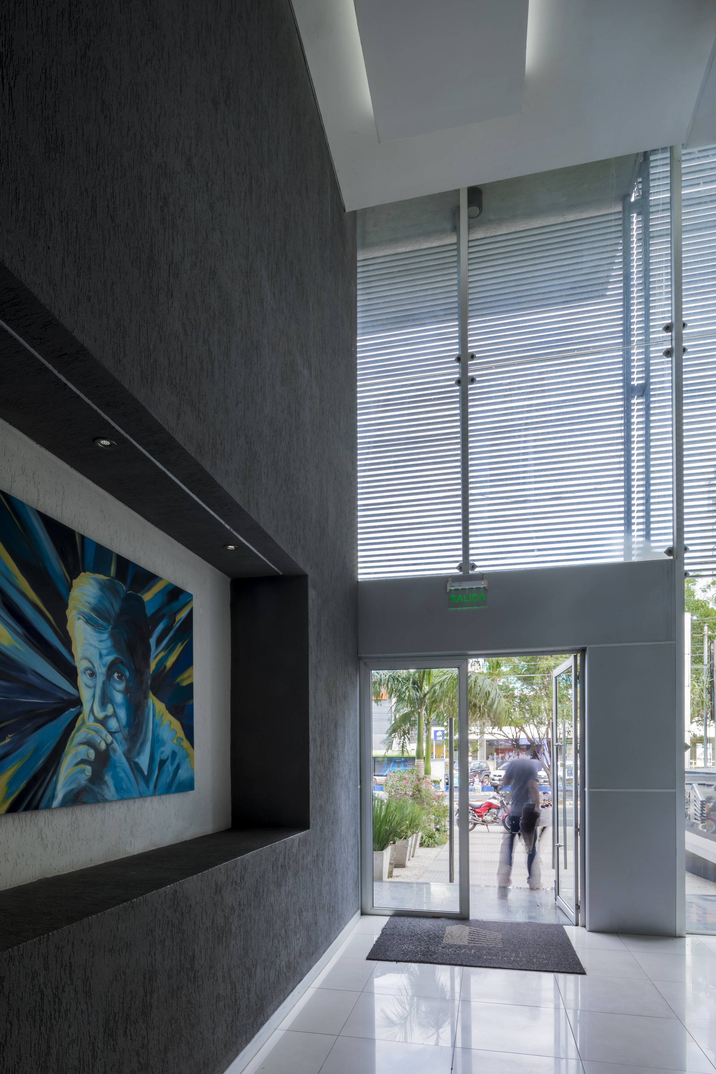 PRO ARQUITECTURA PARAGUAY architecture design PROSA
Interior de un edificio con una puerta de vidrio, un mural con la imagen de una persona pensativa y cortinas de alambre en las ventanas