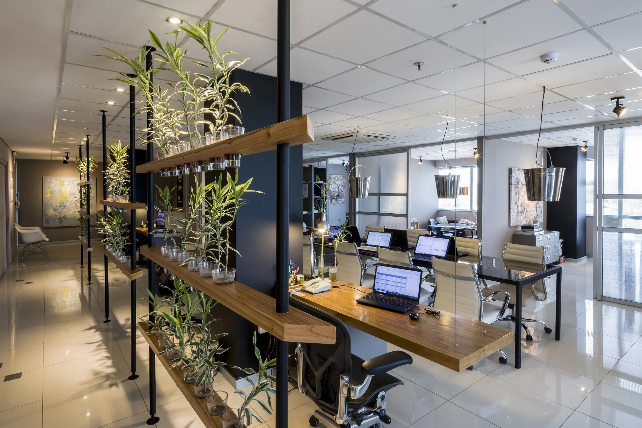 PRO ARQUITECTURA PARAGUAY architecture design PROSA
Oficina moderna con escritorios de madera, sillas blancas, plantas en niveles y lámparas metálicas colgantes, paredes decoradas con arte y ventanas grandes con vista al centro urbano.