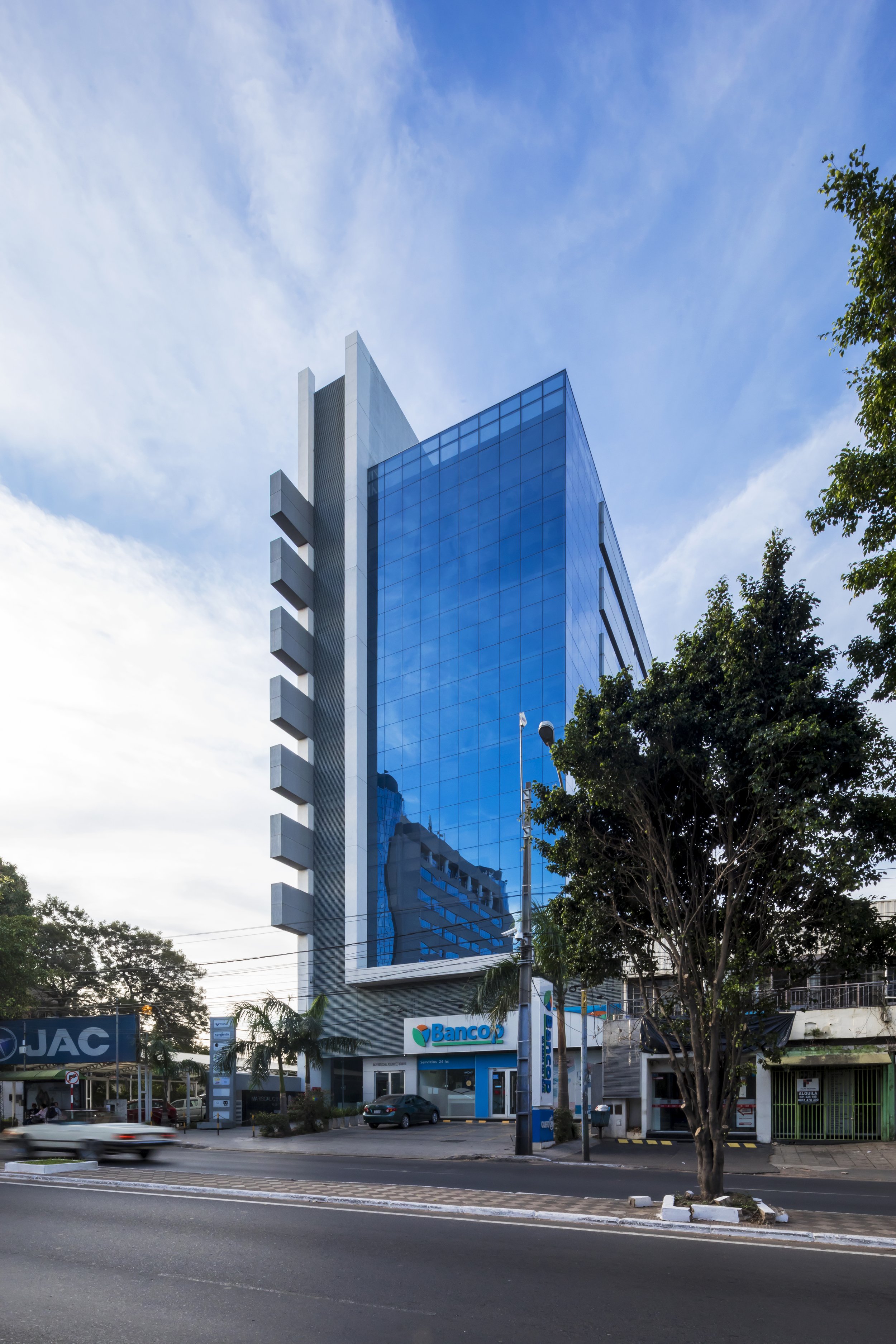 Edificio moderno de vidrio azul en una ciudad, reflejando otros edificios y el cielo. En la base hay un banco de bank y árboles en la calle.