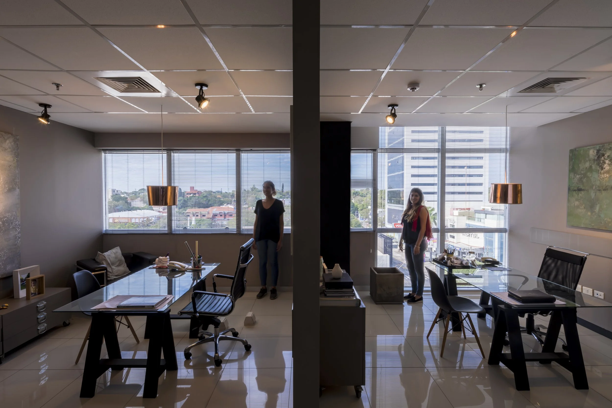 PRO ARQUITECTURA PARAGUAY architecture design PROSA
Salón de oficina con dos mujeres, una en cada lado, con vista a la ciudad a través de grandes ventanas con persianas. Muebles de oficina y objetos en las mesas.