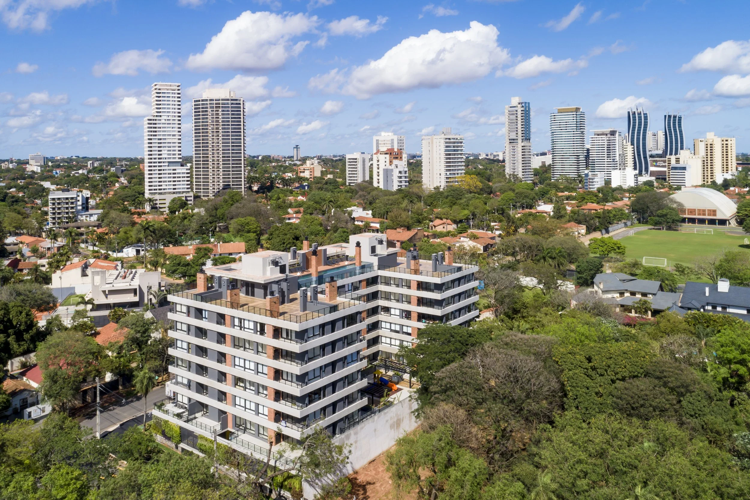 Tour de edificios altos y rascacielos en una ciudad moderna con áreas verdes y casas residenciales cercanas, bajo un cielo soleado con algunas nubes.