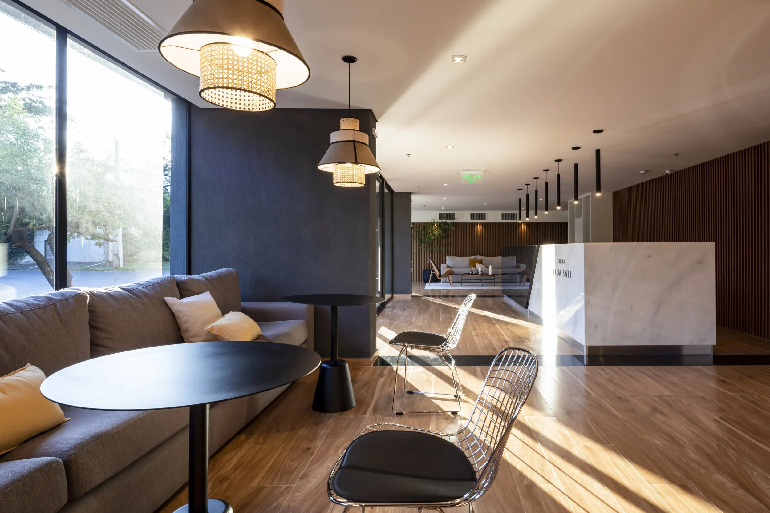 Interior de un vestíbulo moderno con sillones, mesas y lámparas, con grandes ventanales y pisos de madera.