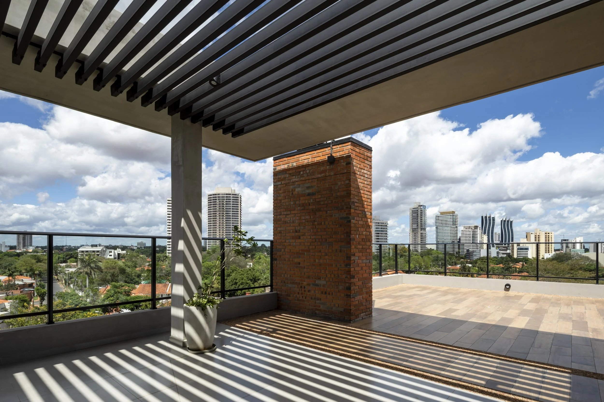 Vista de una terraza con sombra y ciudad de fondo, con edificios altos y cielo con nubes.