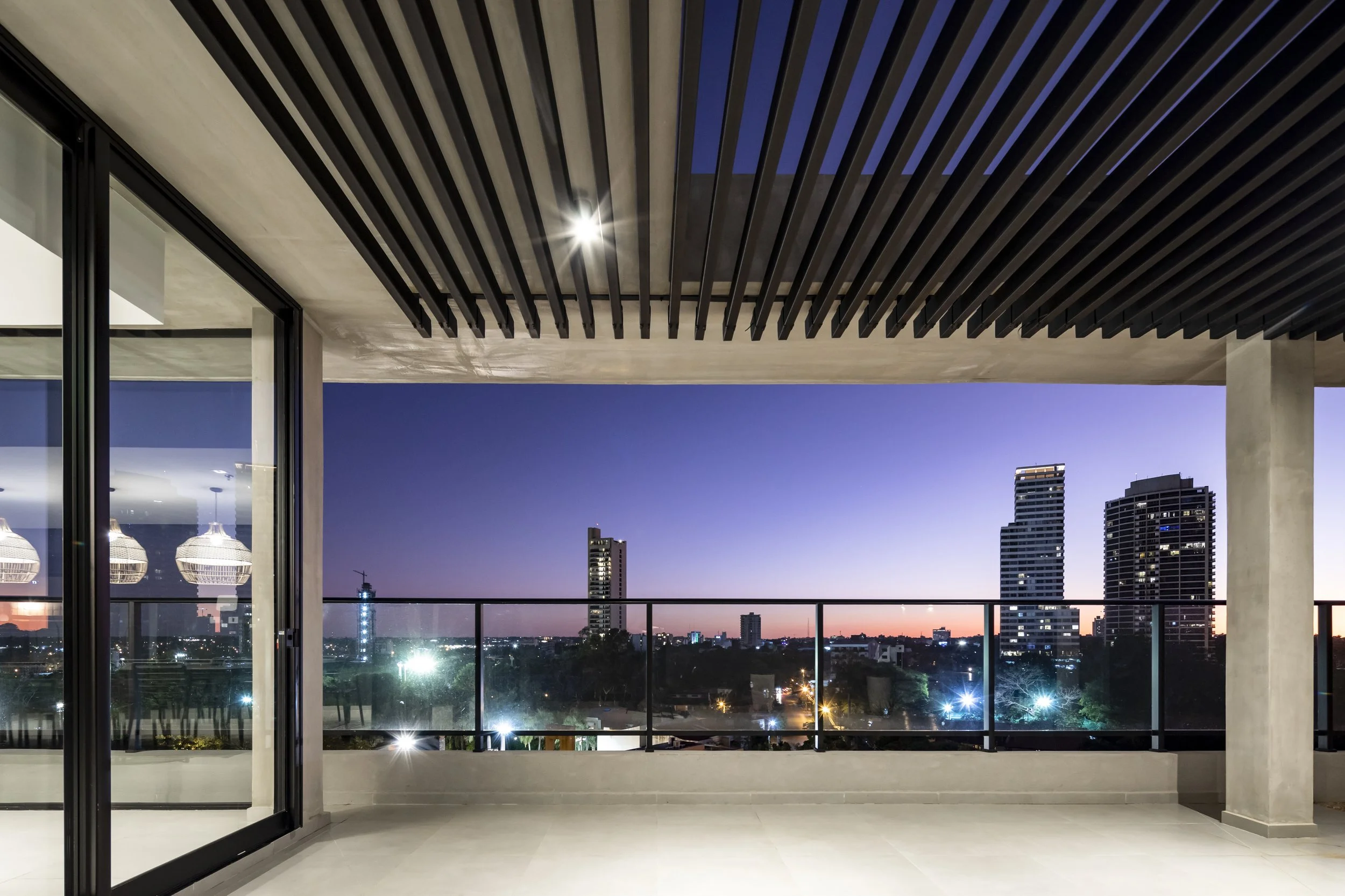 Vista de una terraza moderna con vista a la ciudad durante el atardecer, con cielo azul y edificios altos iluminados.