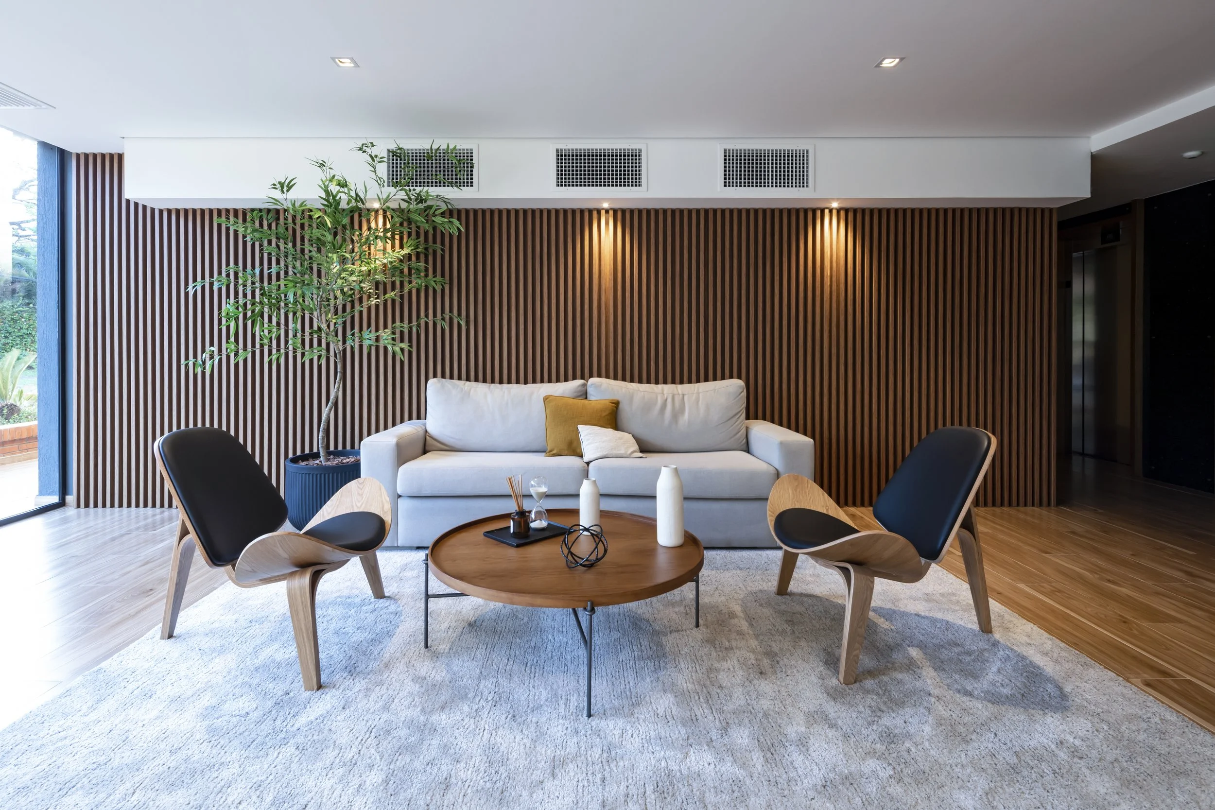 Sala moderna con paredes de panel de madera, sillón blanco con cojín amarillo, mesa redonda de madera, dos sillones con asiento de madera y respaldo negro, planta en maceta azul, zona iluminada con lámparas de techo, alfombra clara y ventanas grandes