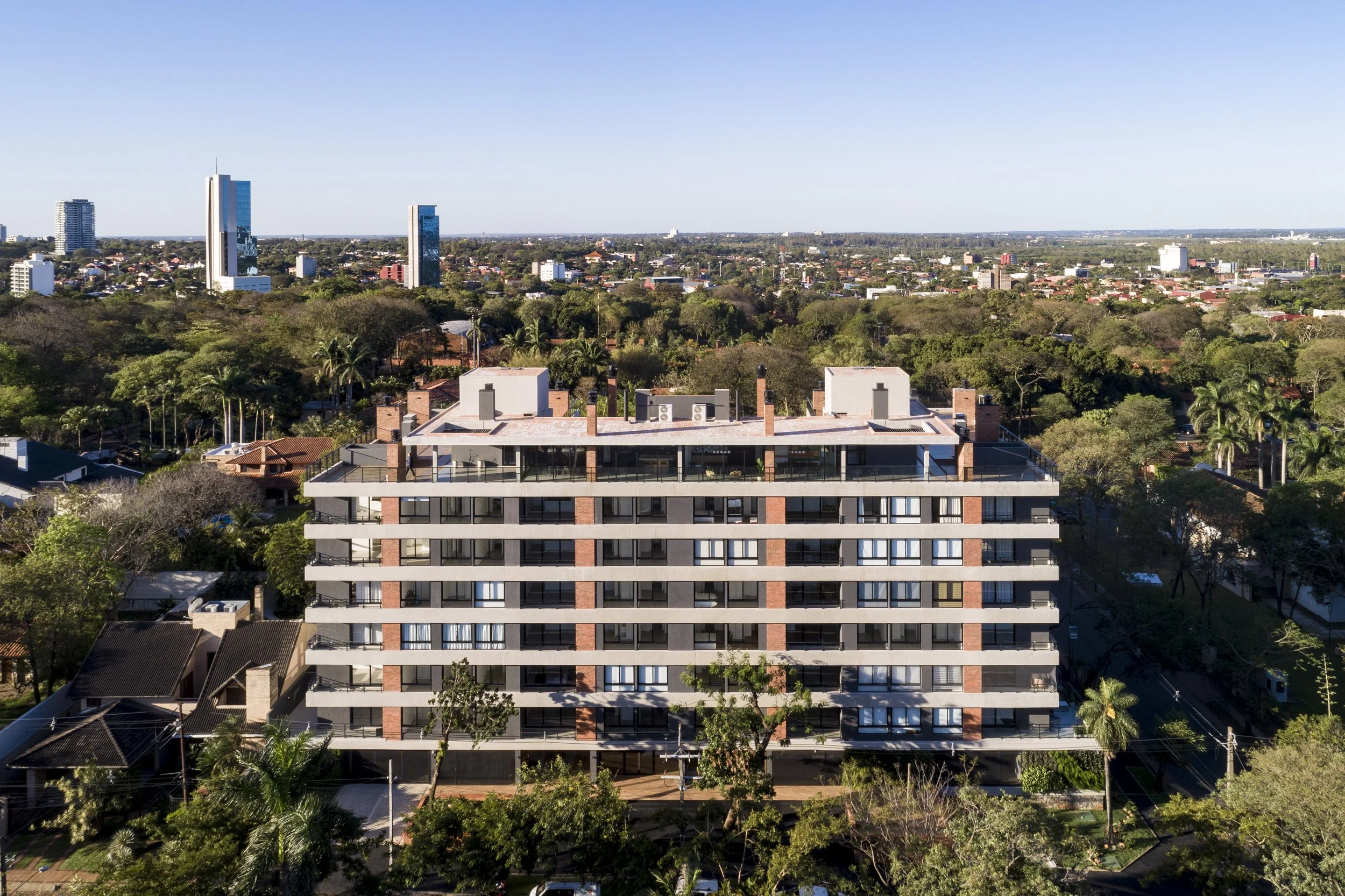 Edificio moderno de varios pisos en un entorno urbano rodeado de árboles y áreas residenciales, con edificios altos en el fondo bajo un cielo despejado.