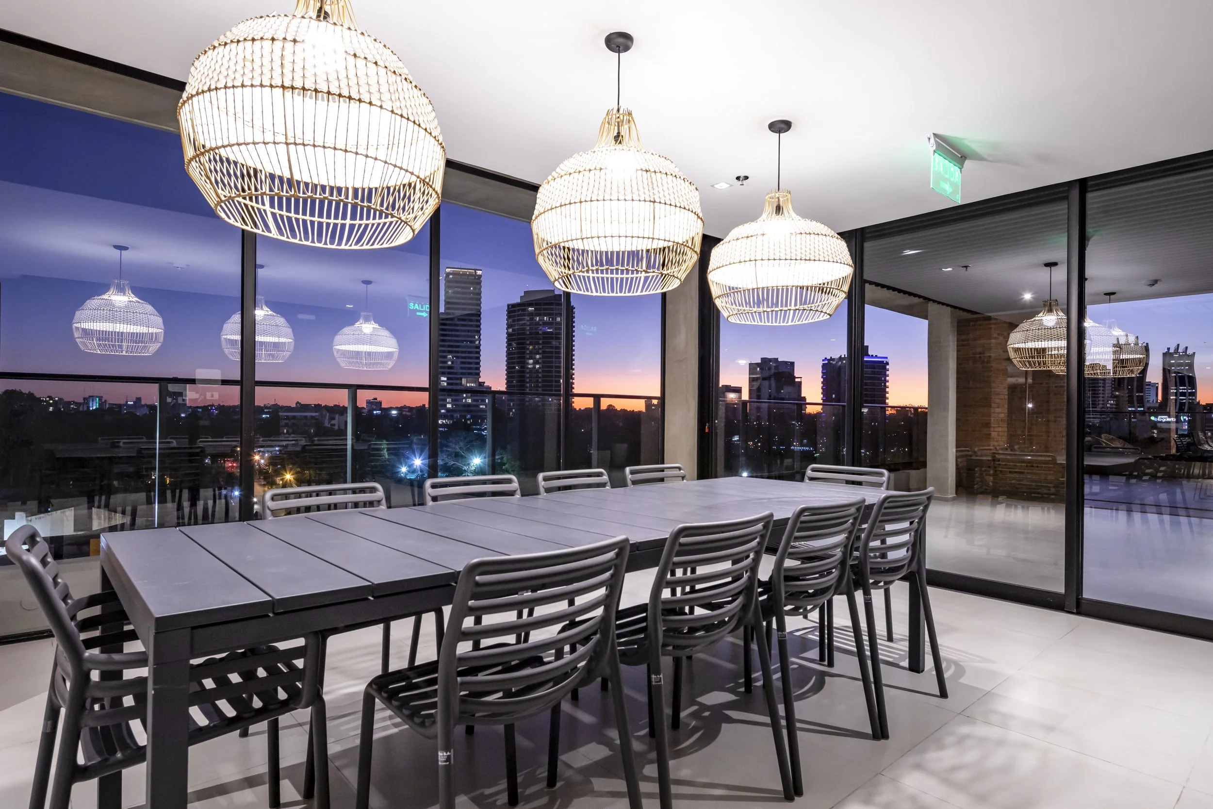 Comedor en un edificio con vista a la ciudad al atardecer, con mesa grande y sillas, luminarias de mimbre colgantes