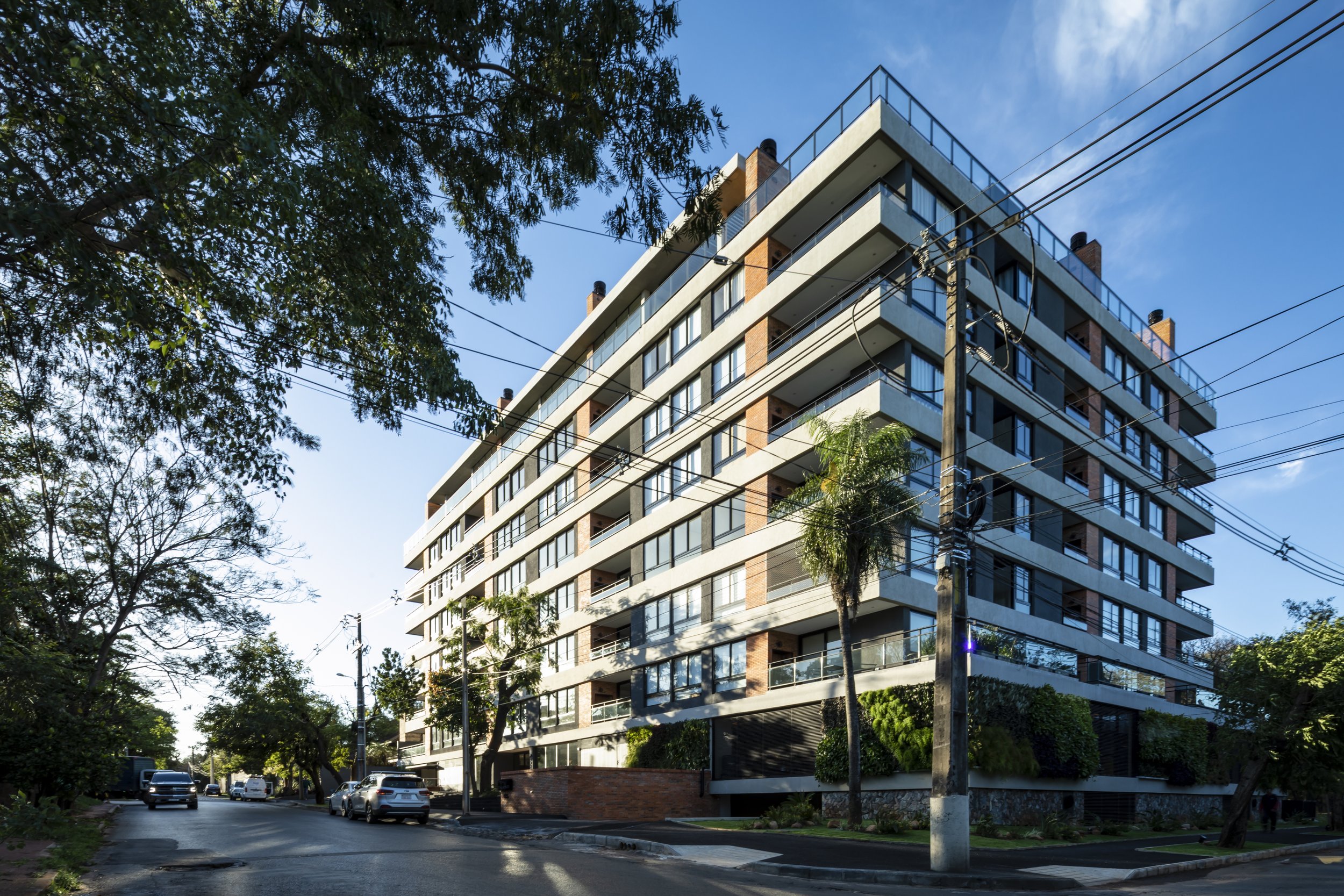 Edificio moderno de varios pisos con balcones y ventanas grandes, ubicado en una calle tranquila con árboles y vehículos estacionados, día soleado.