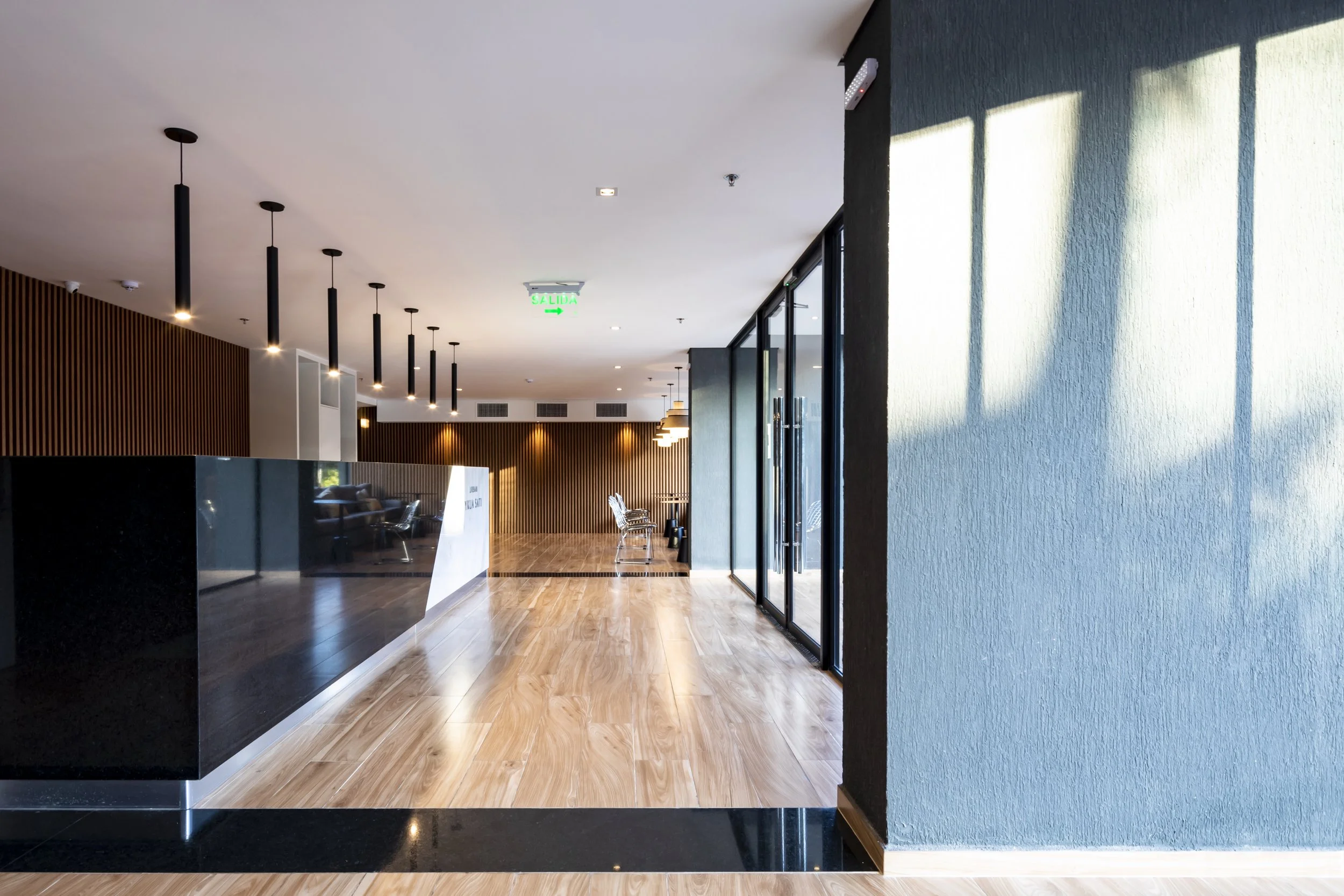 Recepción moderna en un vestíbulo con pisos de madera, paredes con paneles de madera y una pared gris. Hay sofás y lámparas de techo en el fondo, y un cartel de salida en la parte superior.