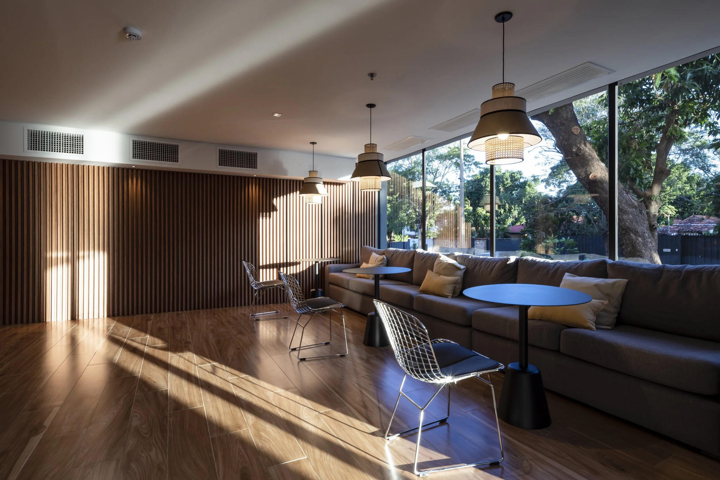 Sala moderna con sillones gris y mesas redondas negras, paredes de madera y ventanas grandes con vista a árboles y naturaleza exterior.