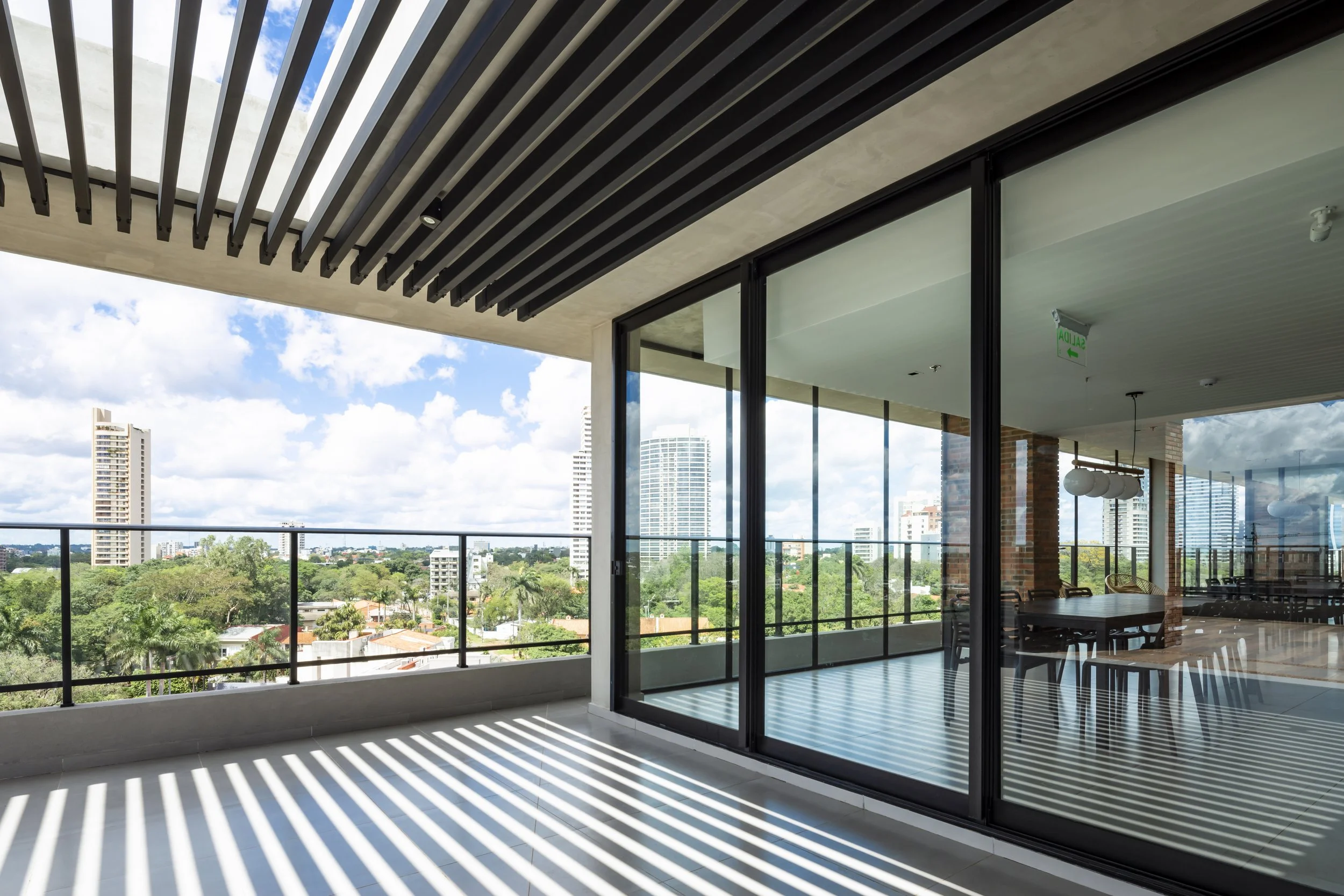 Vista de un balcón con barandal de metal, piso claro, techo con vigas oscuras y vista de edificios y árboles verdes, con puertas de vidrio que llevan a una sala interior con mesa y sillas, en un día soleado con cielo nublado.
