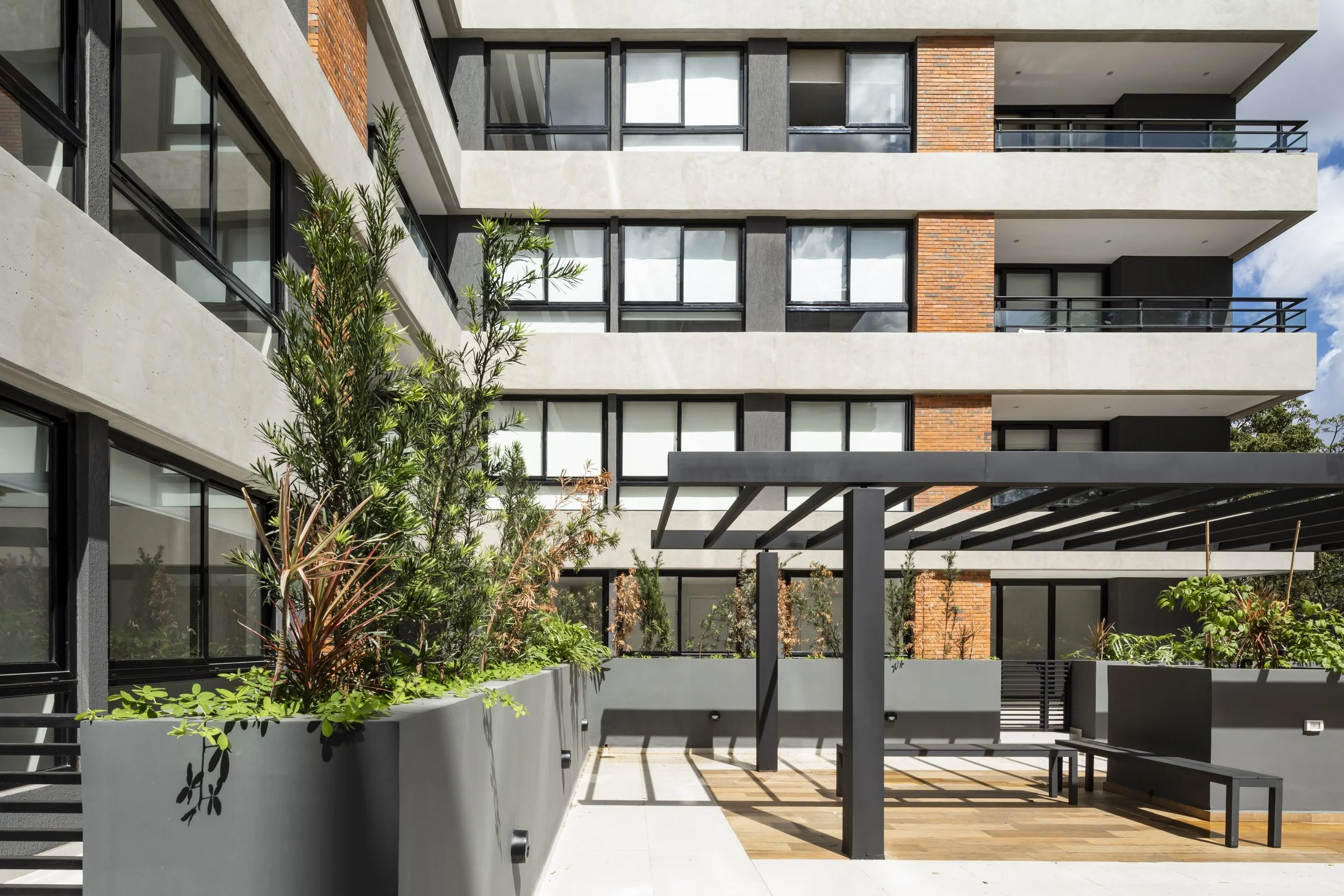 Vista exterior de un edificio moderno de varios pisos con balcones y ventanas grandes, con área de jardín y banca en primer plano.
