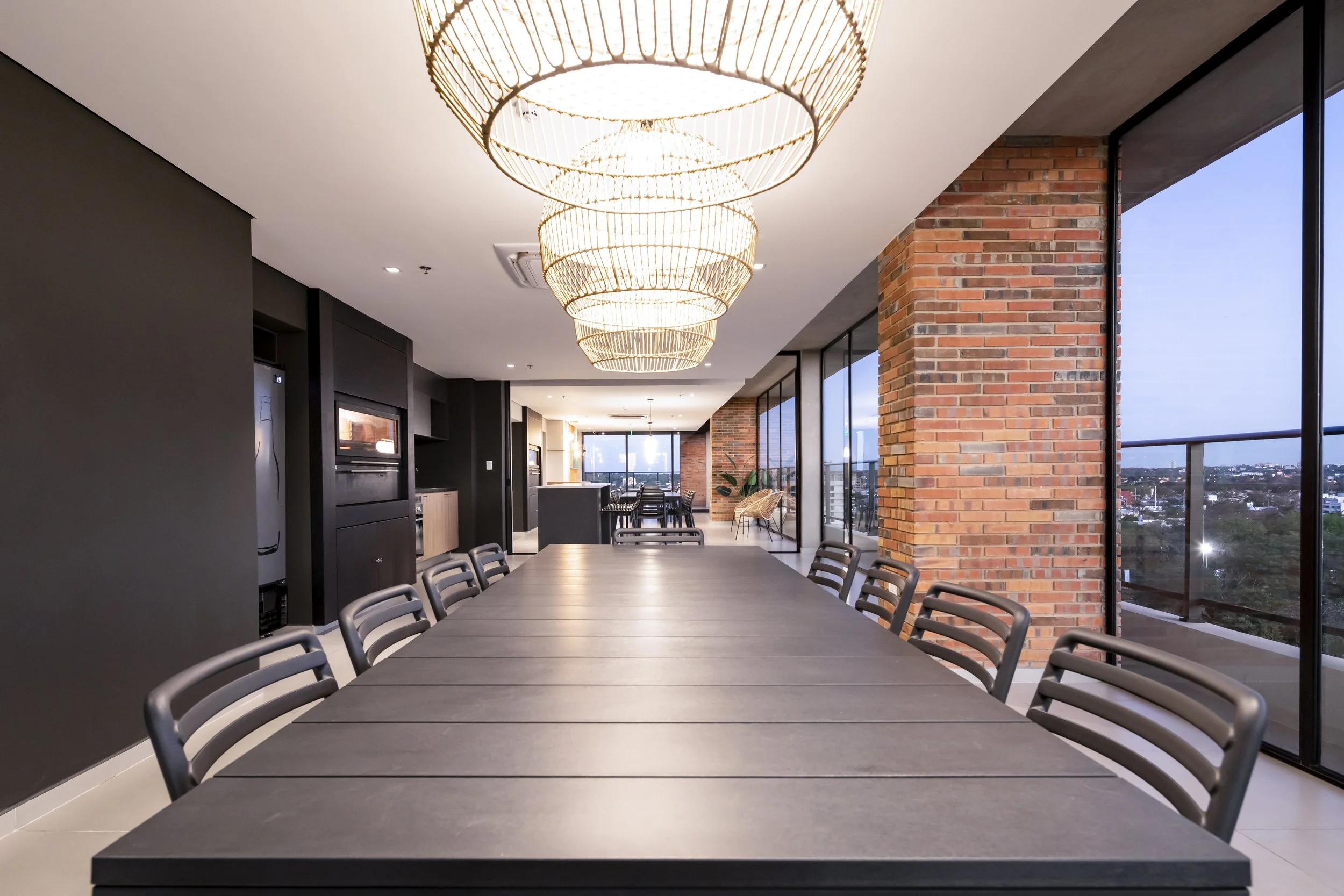 Sala de reuniones moderna con mesa larga, sillas negras, paredes de ladrillo y ventanales grandes con vista a la ciudad, decorada con lámparas de mimbre suspendidas.