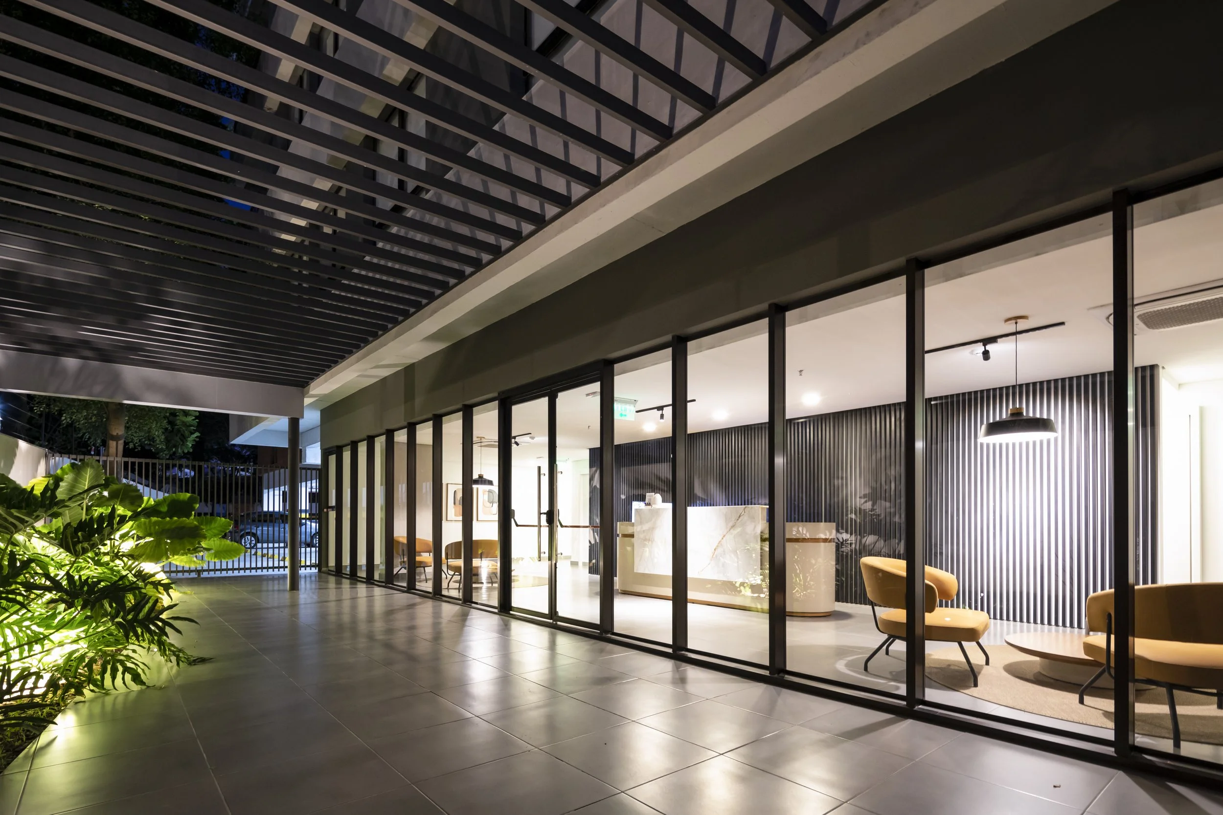 	
PRO ARQUITECTURA PARAGUAY architecture design PROSA
Vista exterior de un lobby de hotel o edificio moderno con paredes de cristal y mobiliario de sala de espera, con plantas al lado y vista hacia la calle en la noche.