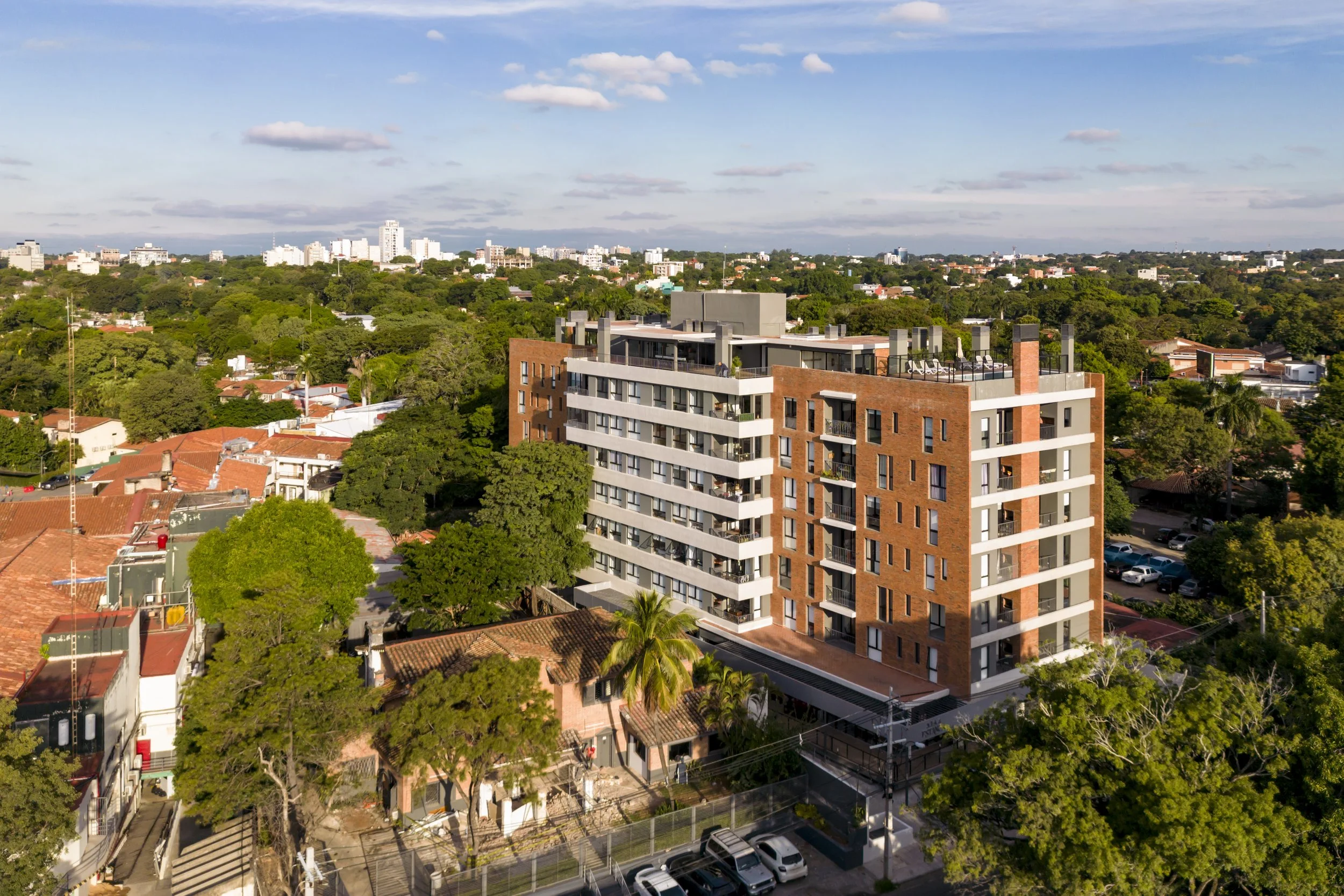 	
PRO ARQUITECTURA PARAGUAY architecture design PROSA
Edificio de apartamentos en un vecindario rodeado de árboles y casas con techos de teja, bajo un cielo parcialmente nublado y un horizonte urbano en la distancia.