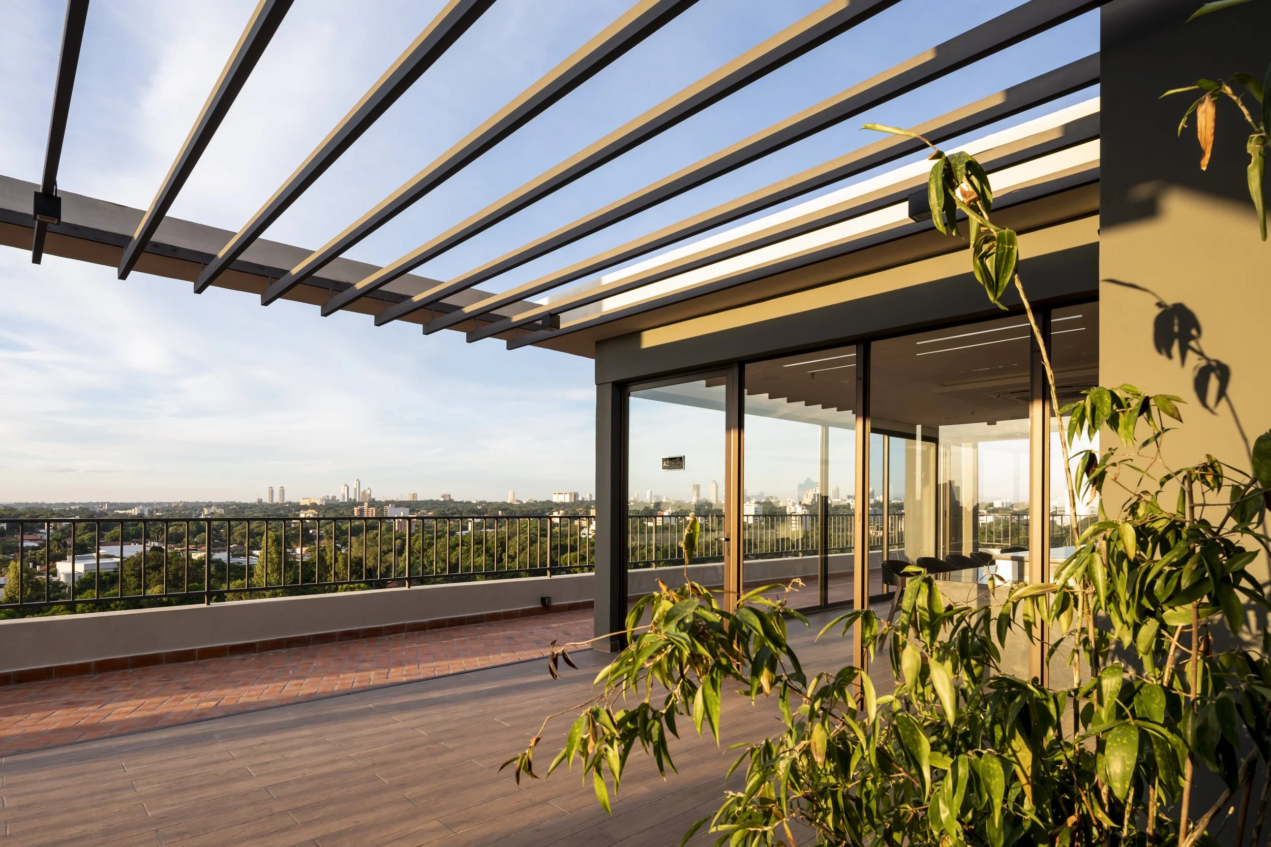 Vista de una terraza en un edificio alto con plantas y vista urbana en el fondo, durante el día.