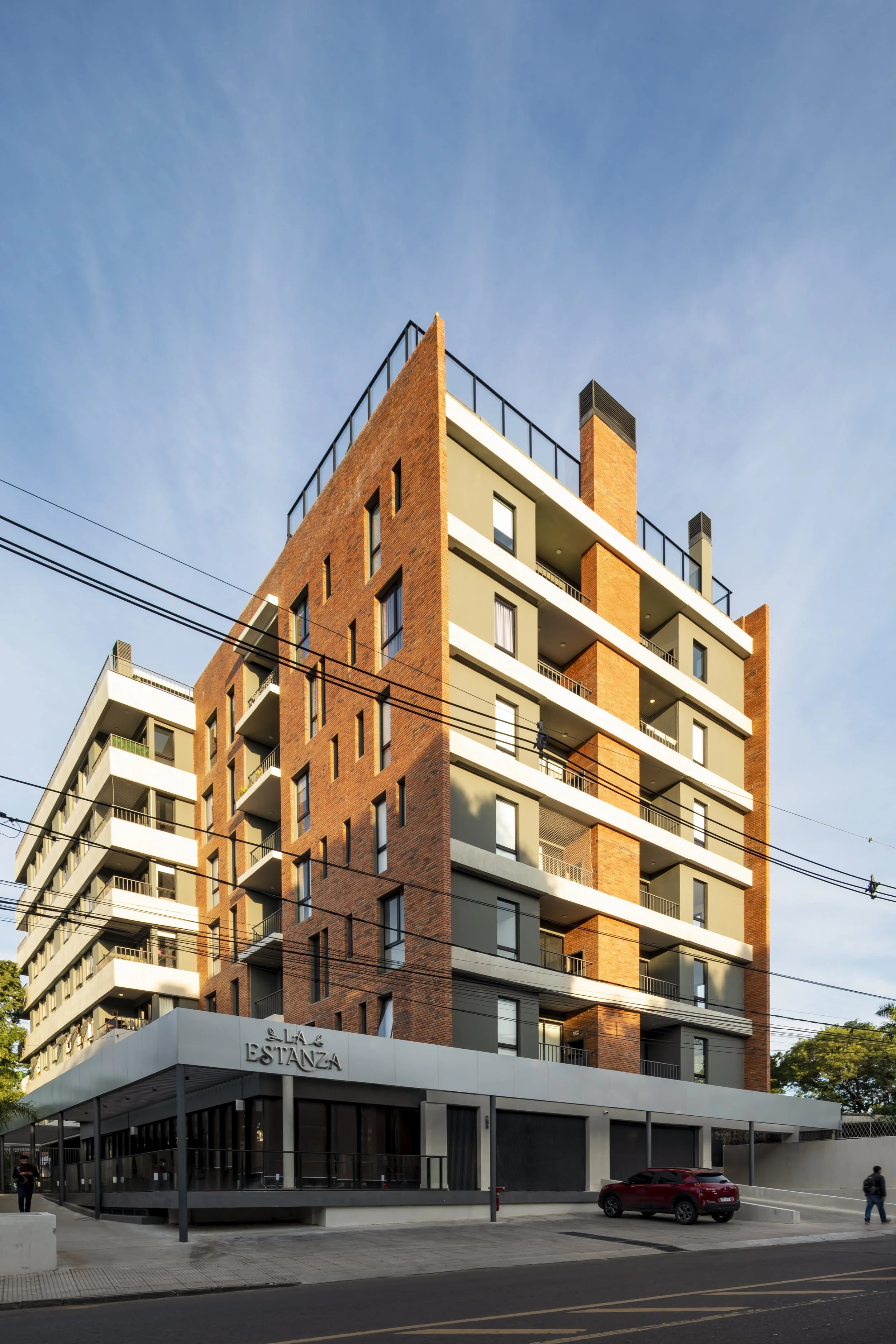 Edificio residencial de varios pisos con fachada de ladrillo y balcones, en la planta baja un local comercial con letrero que dice 'La Estanza' y un coche estacionado en la calle