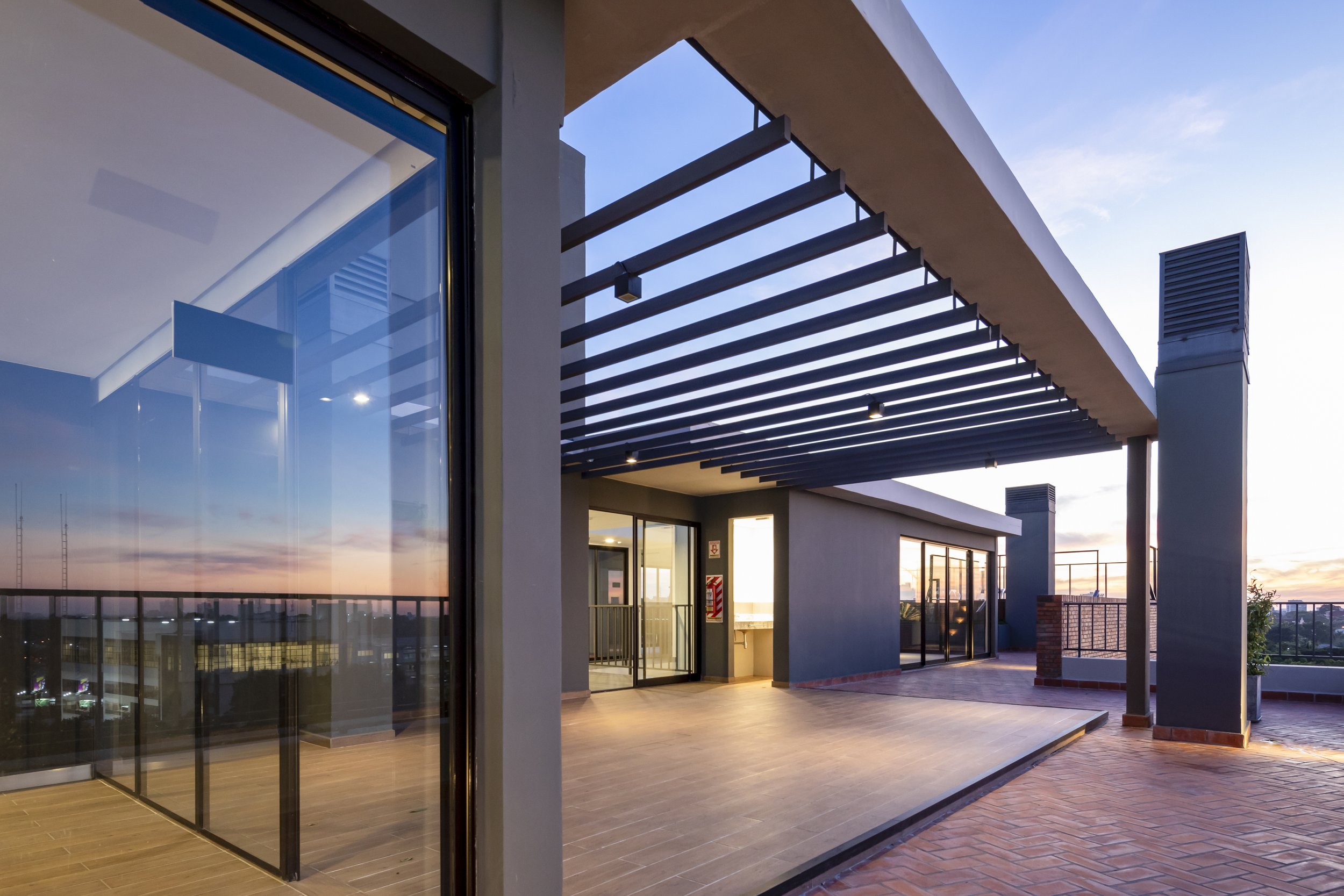 	
PRO ARQUITECTURA PARAGUAY architecture design PROSA
Terraza moderna en atardecer, con piso de ladrillo, paredes grises, techos con vigas negras y ventanas de vidrio, vista a la ciudad y cielo con nubes.