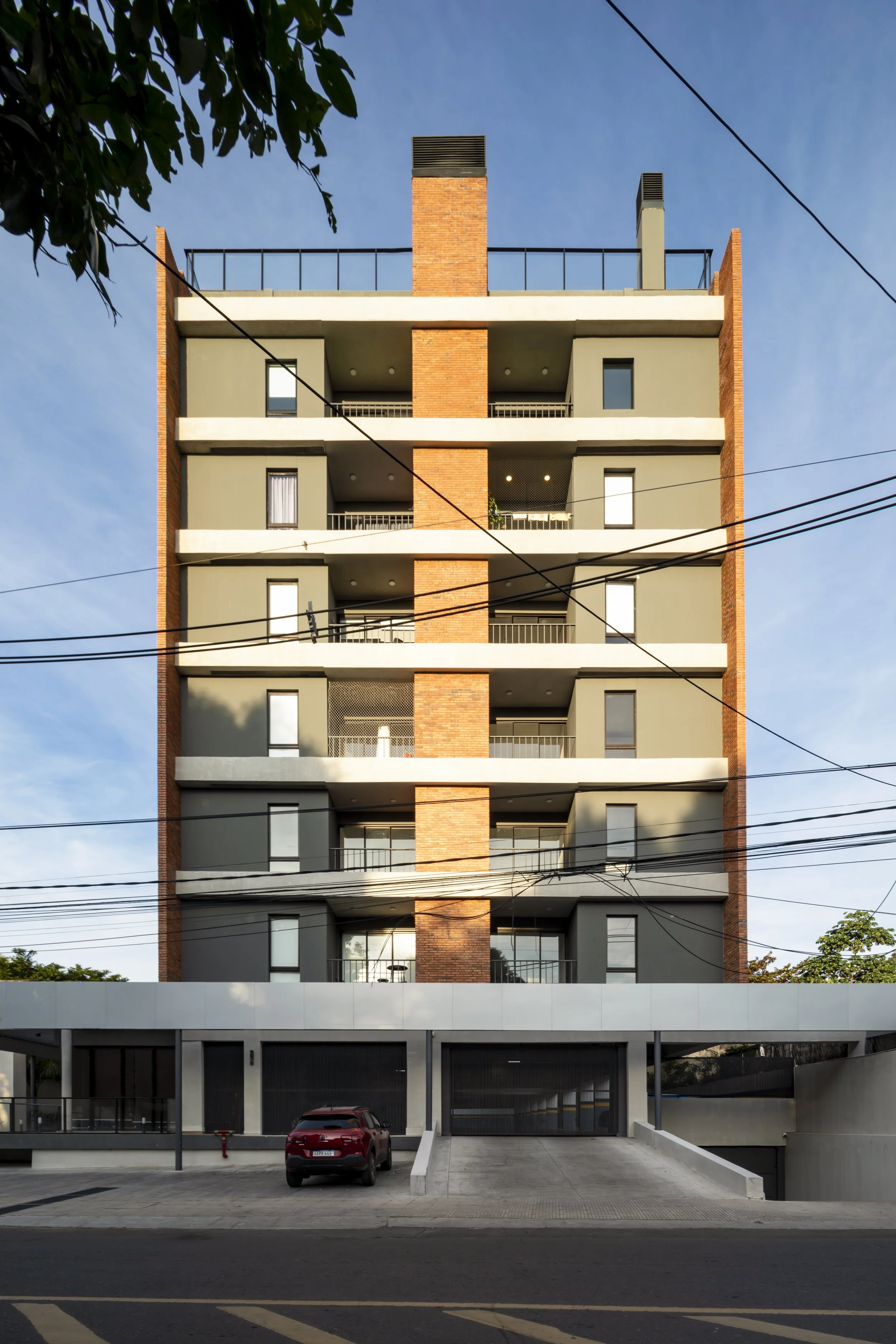 Edificio de apartamentos de varios pisos con fachada moderna y balcones, con un coche estacionado en la entrada y cables eléctricos cruzando la imagen.