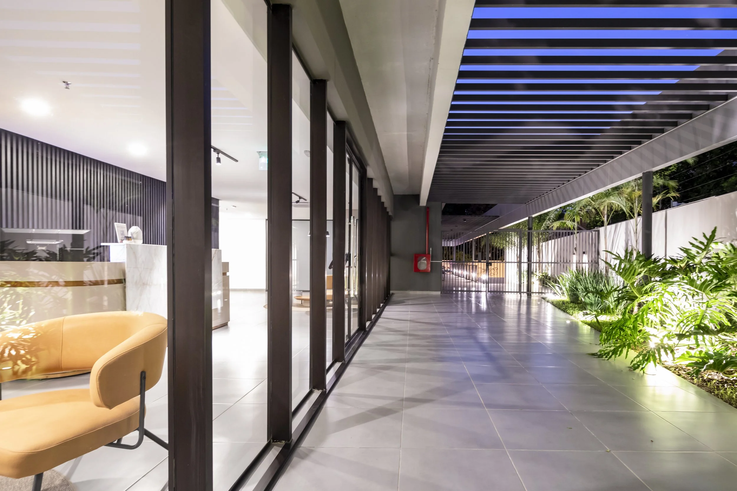 	
PRO ARQUITECTURA PARAGUAY architecture design PROSA
Pasillo exterior de un edificio moderno con plantas y una cerca, vista hacia la noche
