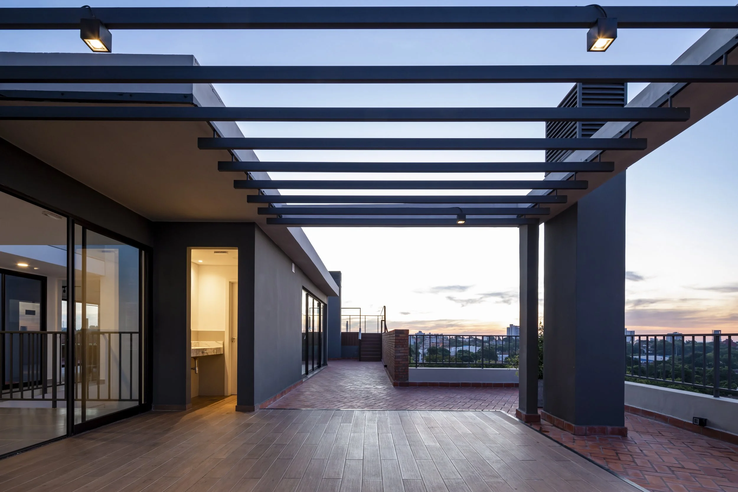 	
PRO ARQUITECTURA PARAGUAY architecture design PROSA
Terraza en un edificio moderno durante el atardecer, con suelo de madera y barandales de metal, vista a la ciudad.