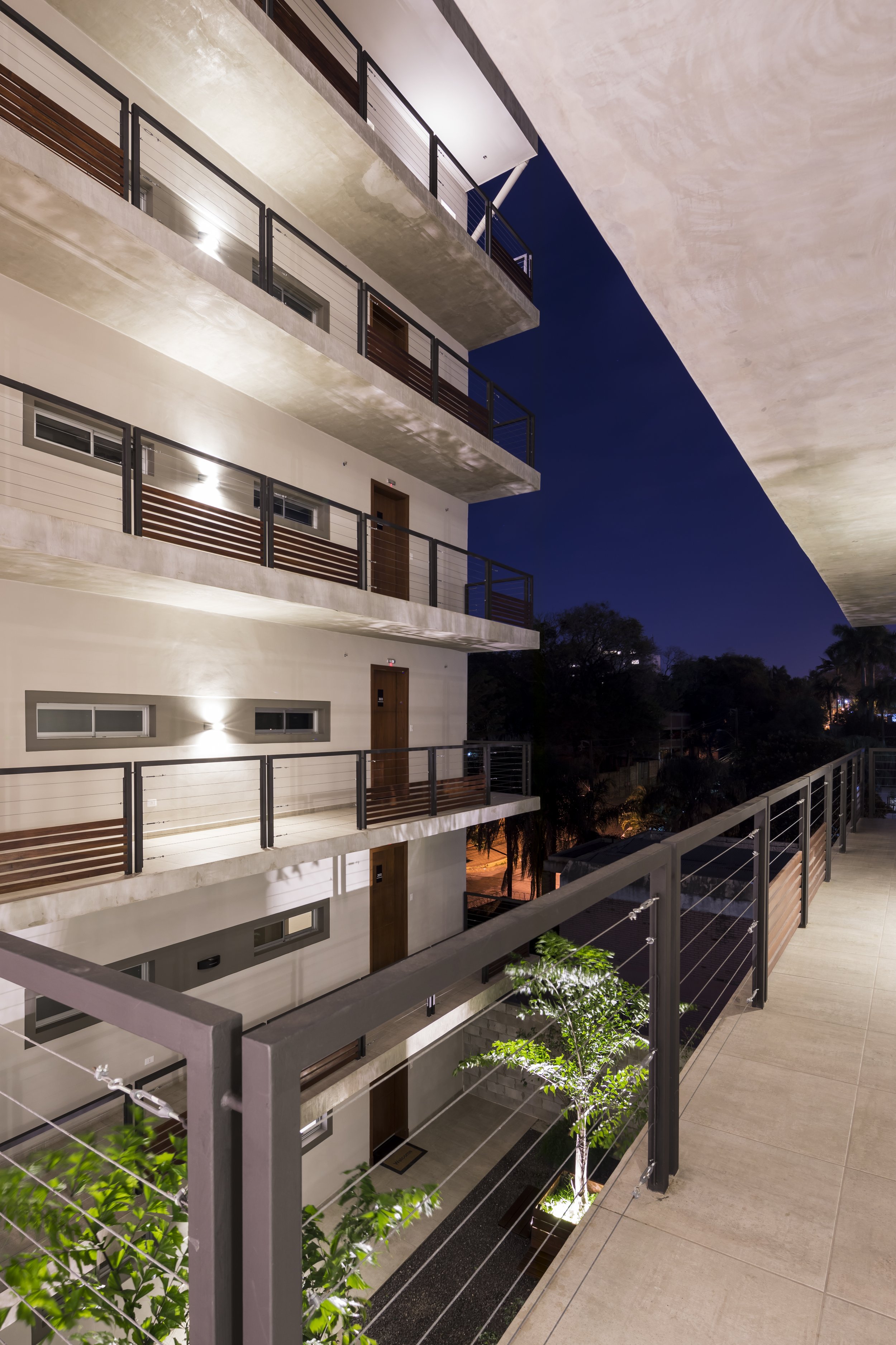 	
PRO ARQUITECTURA PARAGUAY architecture design PROSA
Vista de un balcón en un edificio de varios pisos durante la noche, con paredes blancas, barandales de metal y madera, y plantas decorativas en el área.