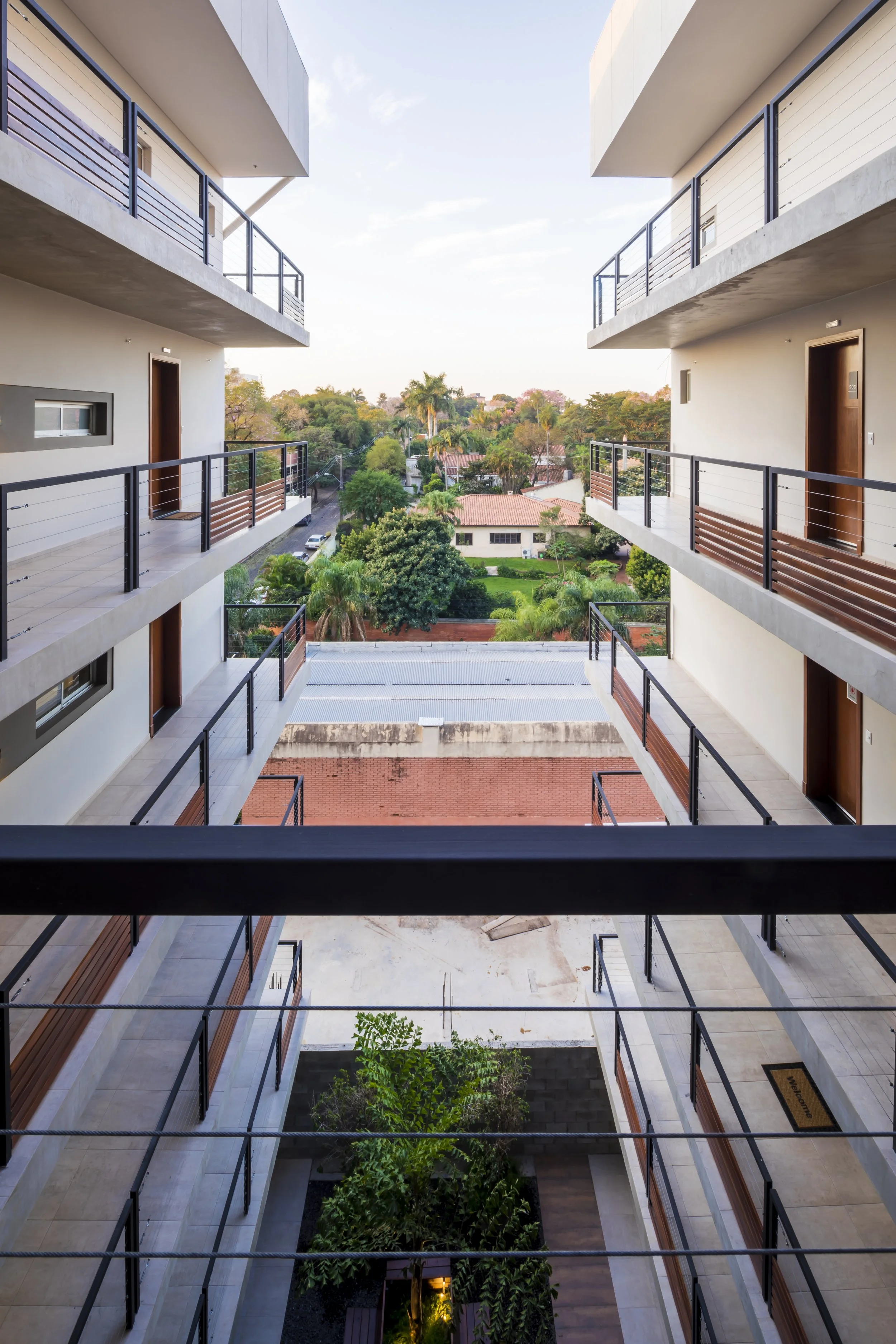 	
PRO ARQUITECTURA PARAGUAY architecture design PROSA
Vista de apartamentos de varios pisos con pasillos y puertas a cada lado, patio interior con árboles y vegetación, y vista de casas y árboles en el fondo bajo un cielo despejado con algunas nubes.