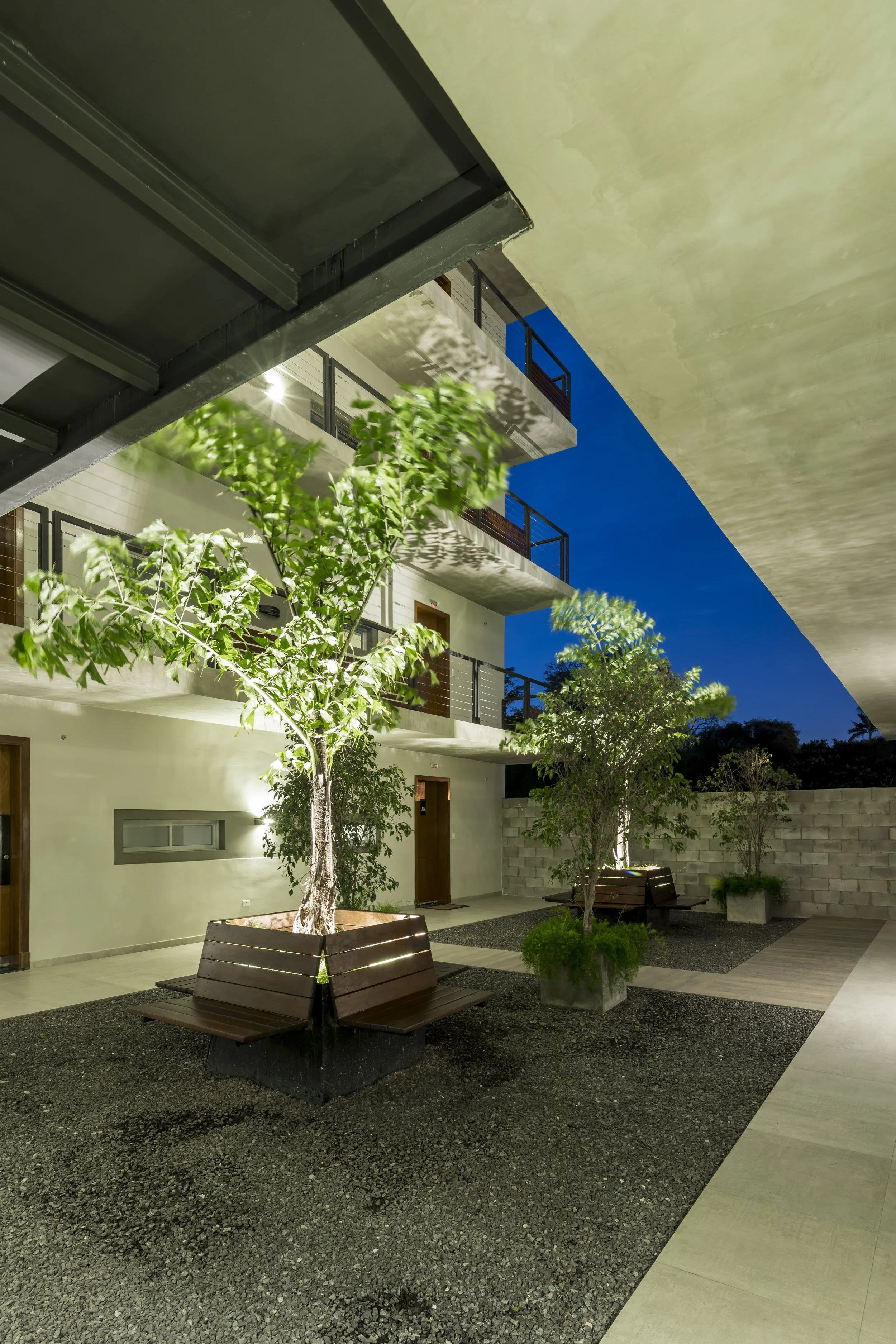 	
PRO ARQUITECTURA PARAGUAY architecture design PROSA
Patio interior de un edificio de varias plantas con árboles en macetas, bancos de madera y paredes de concreto, iluminado por luces artificiales durante la noche.