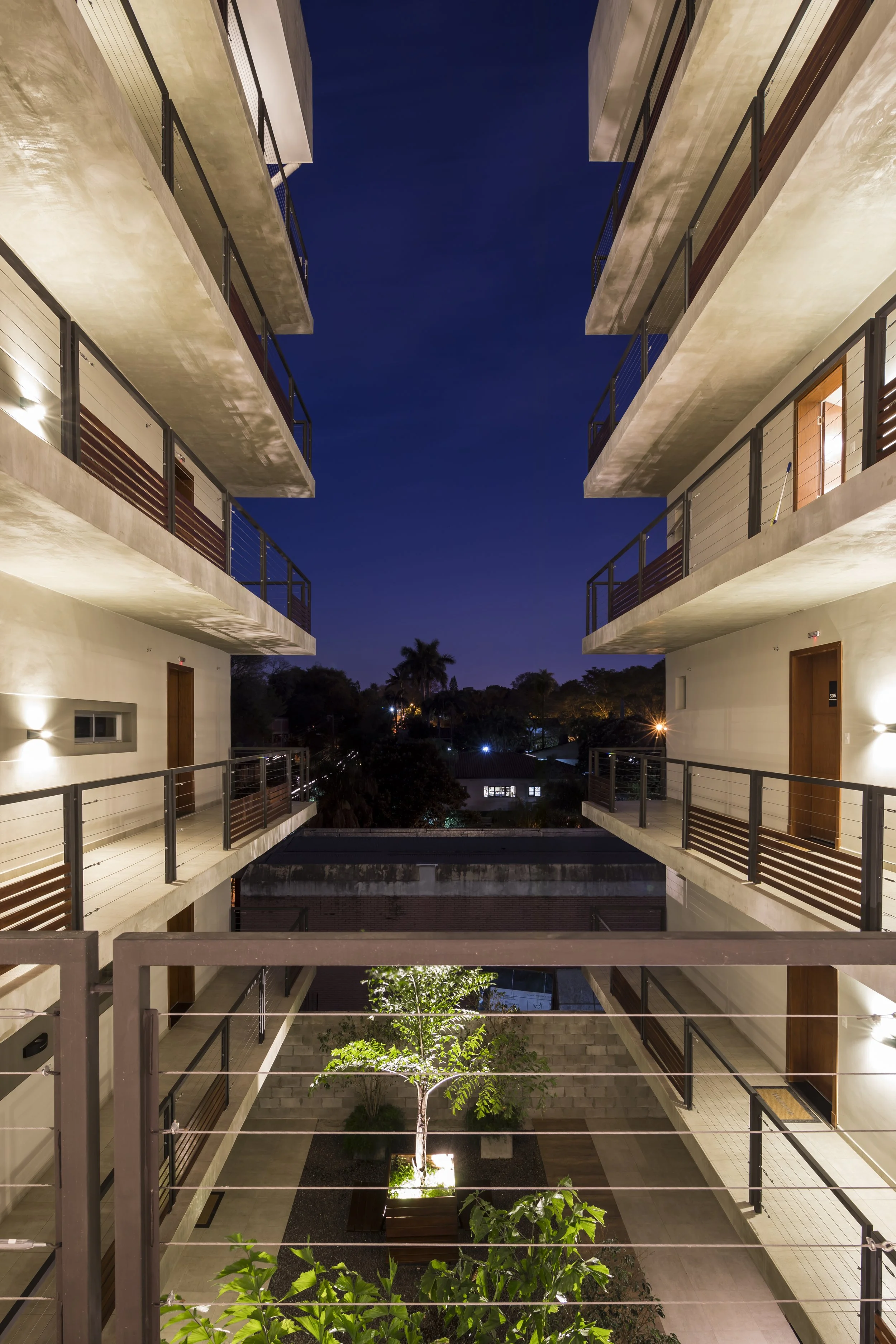 	
PRO ARQUITECTURA PARAGUAY architecture design PROSA
Imagen nocturna de un edificio de apartamentos con pasillos y balcones, y un área interior con un árbol iluminado.
