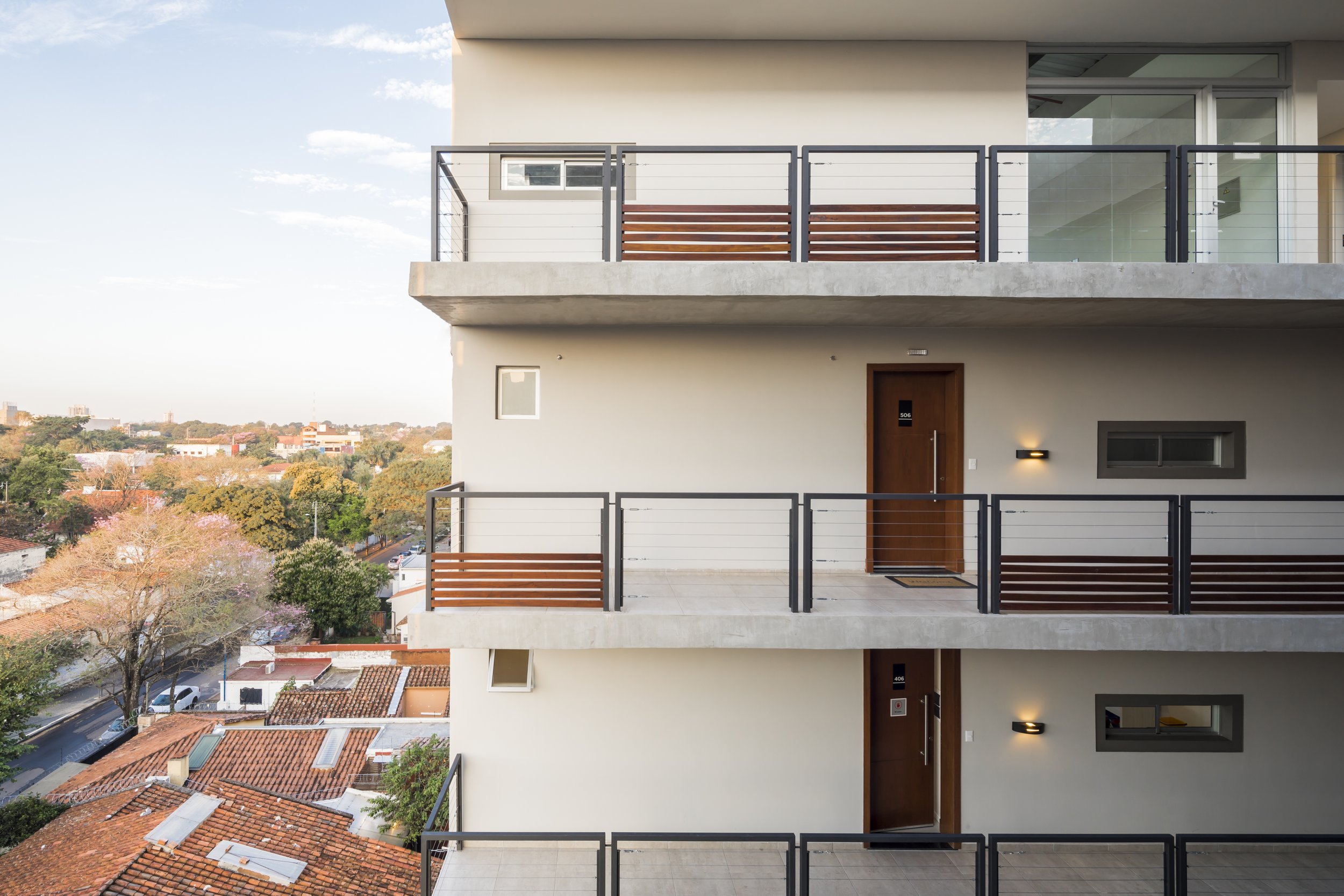 	
PRO ARQUITECTURA PARAGUAY architecture design PROSA
Vista de un edificio moderno de varios pisos con balcones y puertas de madera, con un paisaje urbano y árboles al fondo, durante el día.