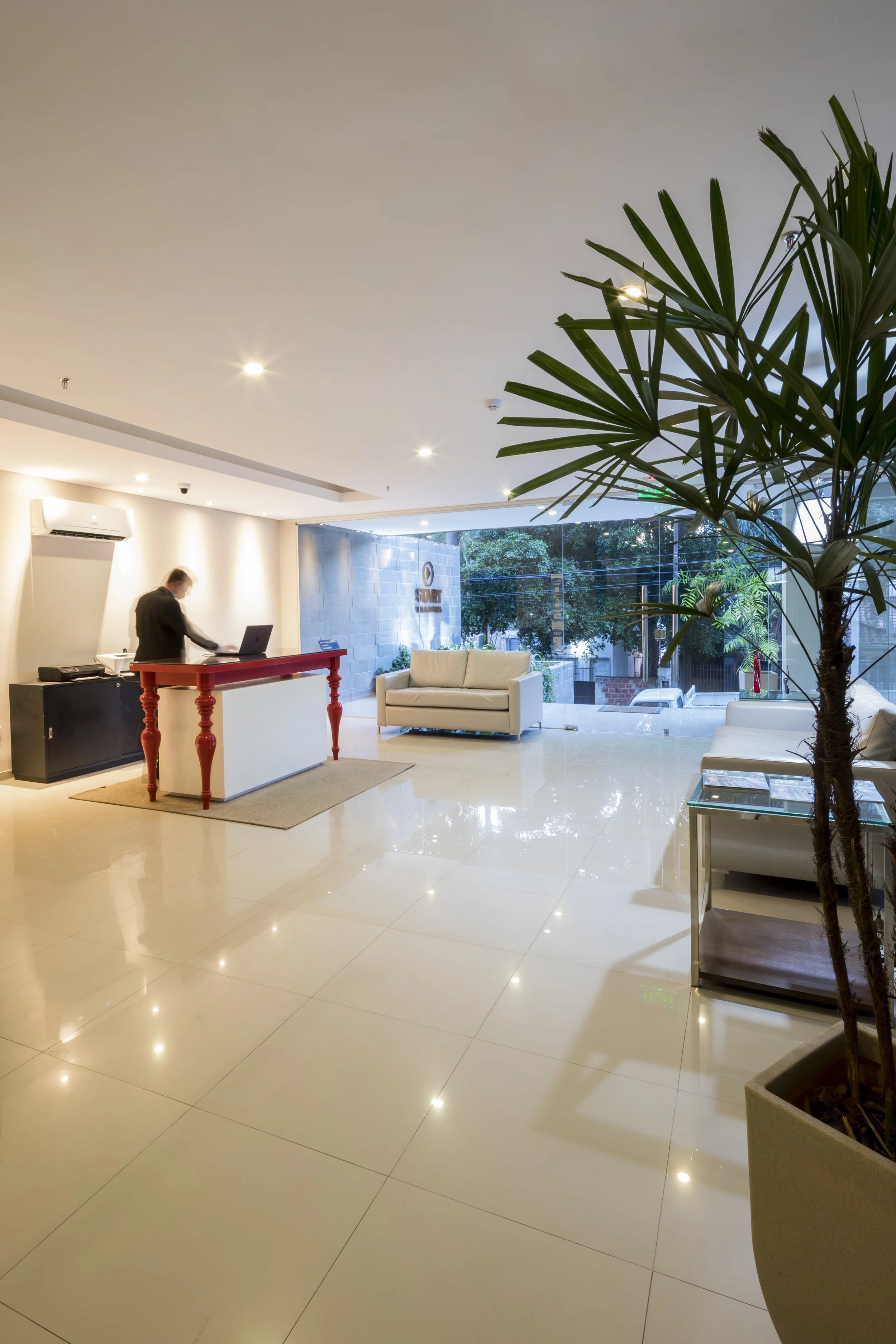 	
PRO ARQUITECTURA PARAGUAY architecture design PROSA
Vestíbulo de un hotel con sillones blancos, una planta grande y recepción con una persona trabajando en una laptop.