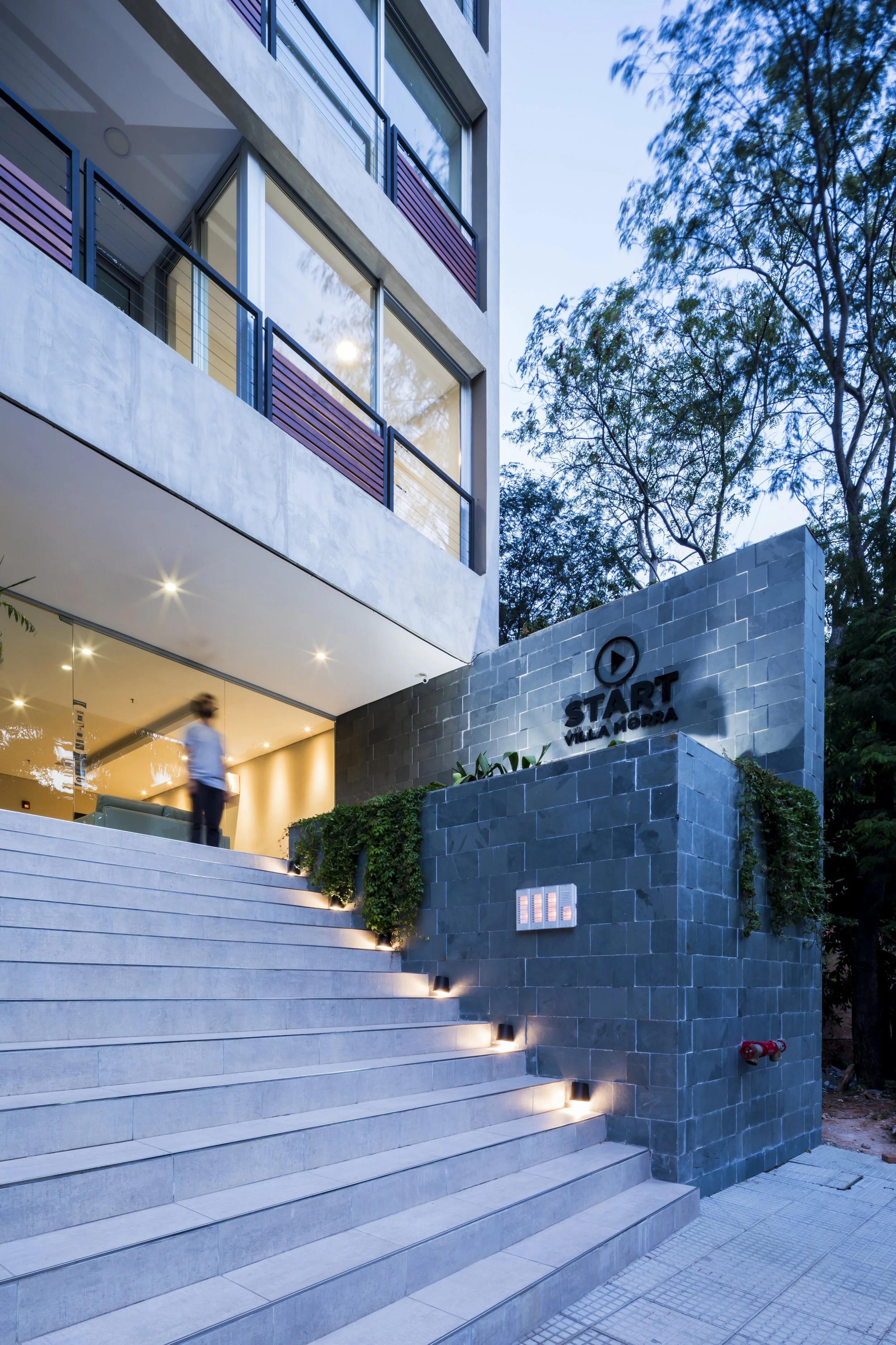 	
PRO ARQUITECTURA PARAGUAY architecture design PROSA
Entrada moderna a una residencia con escaleras iluminadas y señal que dice 'START VILLA MORENA', con un edificio de estilo contemporáneo y árboles en el fondo.