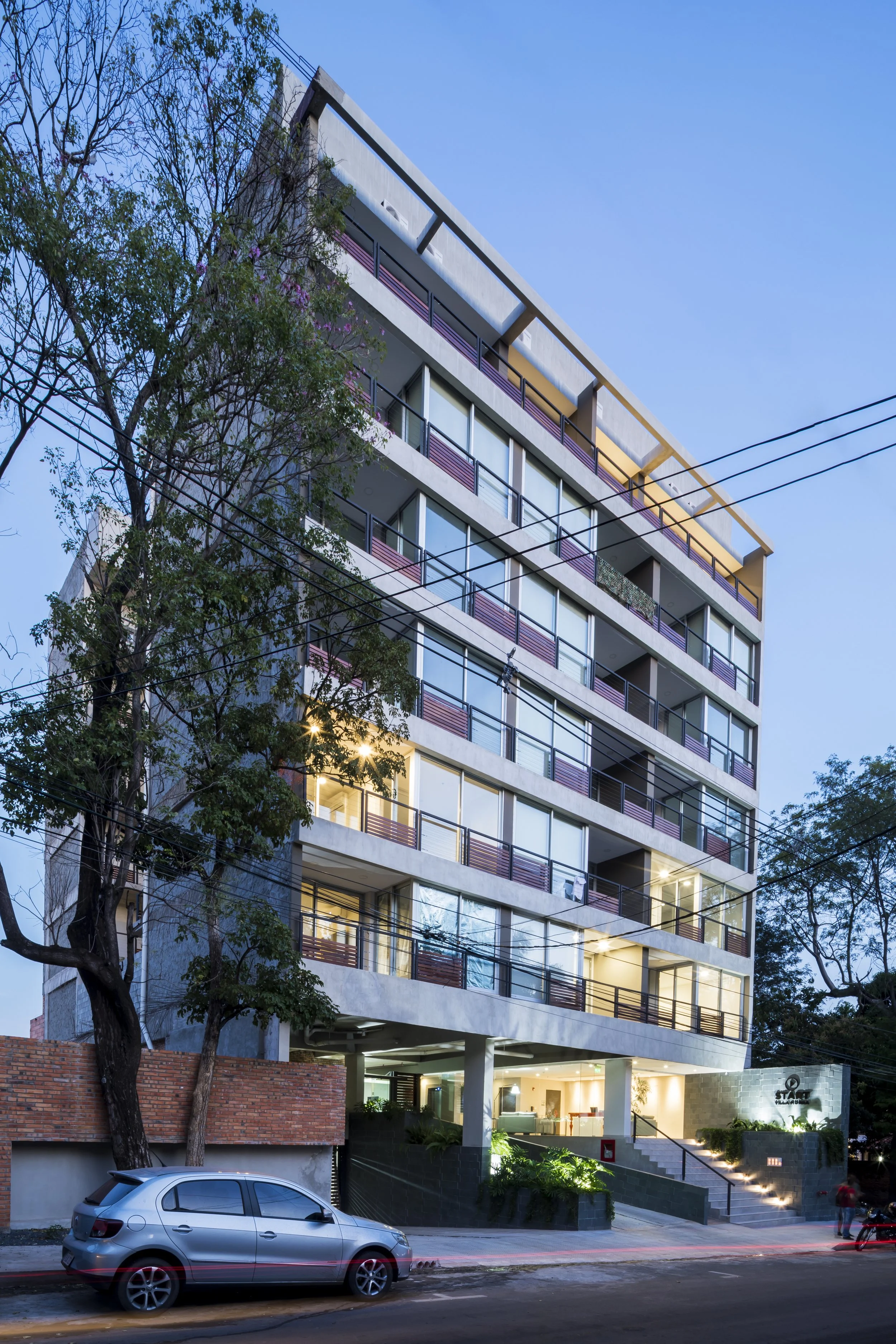 	
PRO ARQUITECTURA PARAGUAY architecture design PROSA
Edificio moderno de varias plantas con ventanas grandes y balcones, luces encendidas en el interior, árboles en la entrada y un coche estacionado frente, en la noche con cielo azul claro.