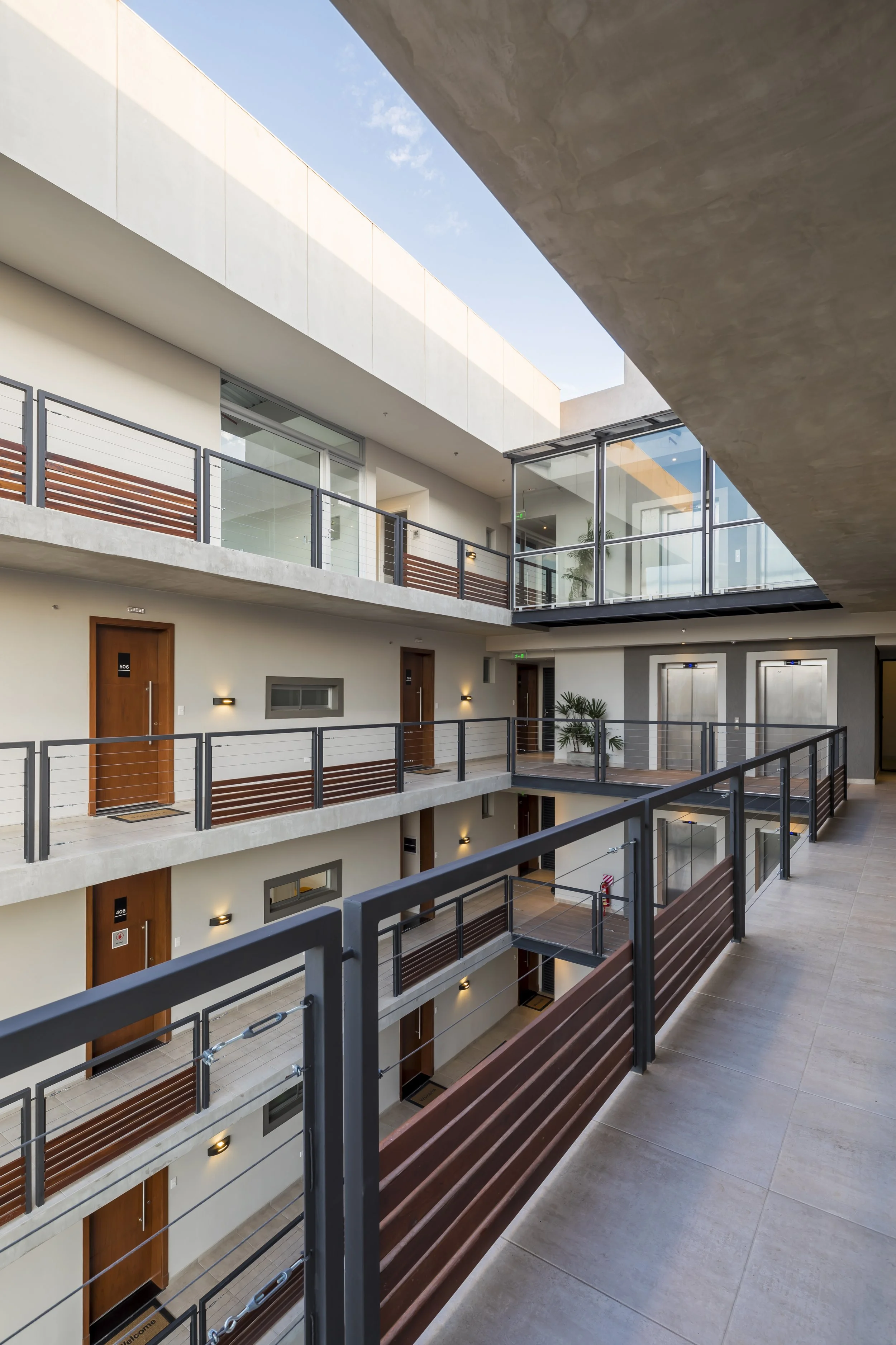 	
PRO ARQUITECTURA PARAGUAY architecture design PROSA
Pasillo interior de un edificio de apartamentos con varias puertas, balcones, barandales de metal negro, paredes blancas y pisos de baldosas beige, con paredes de vidrio y plantas decorativas.