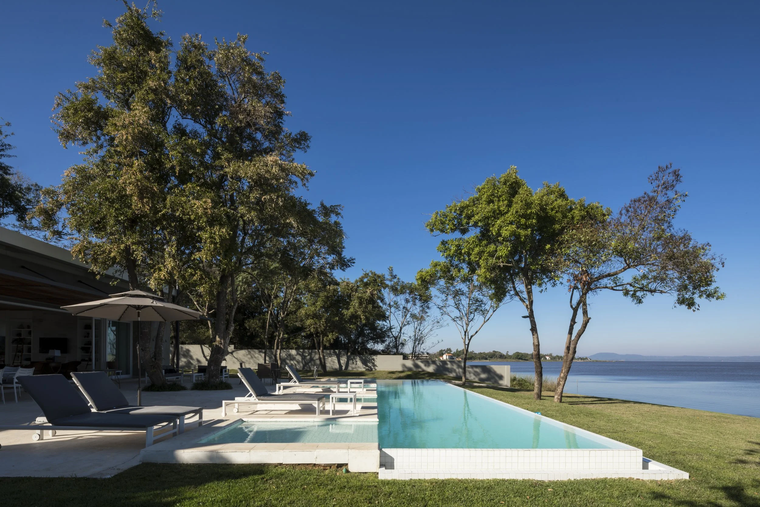 Piscina en un patio con sombras de árboles, sillones y vista al mar en un día soleado.