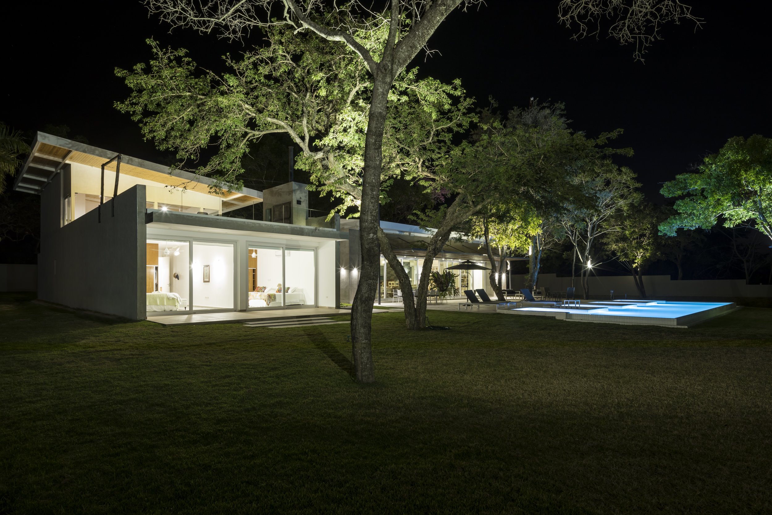 Casa moderna iluminada en la noche con piscina y árboles en el jardín.