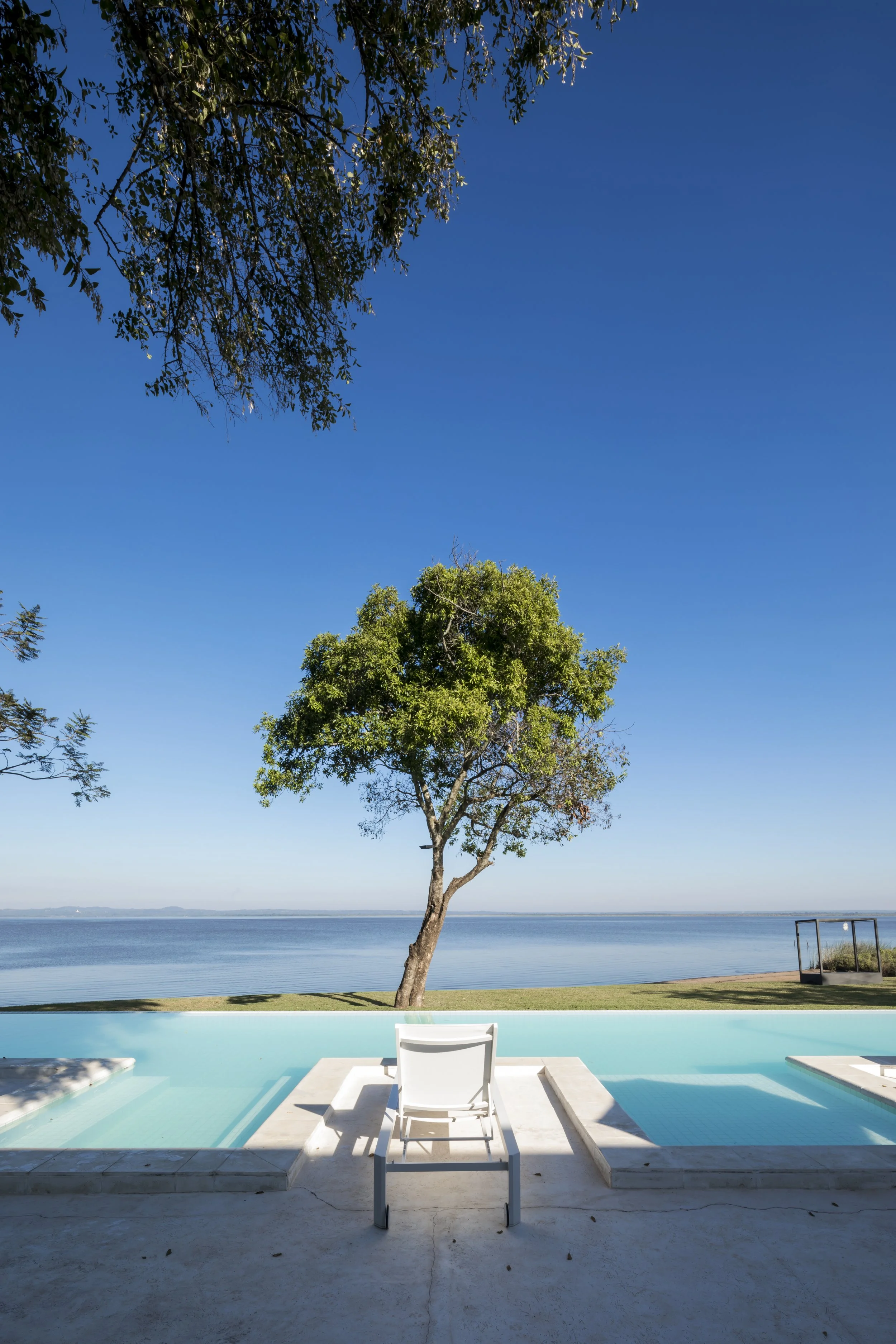	
PRO ARQUITECTURA PARAGUAY architecture design PROSA
Vista de una piscina junto a un lago con un árbol y un sillón blanco en un día soleado y despejado.