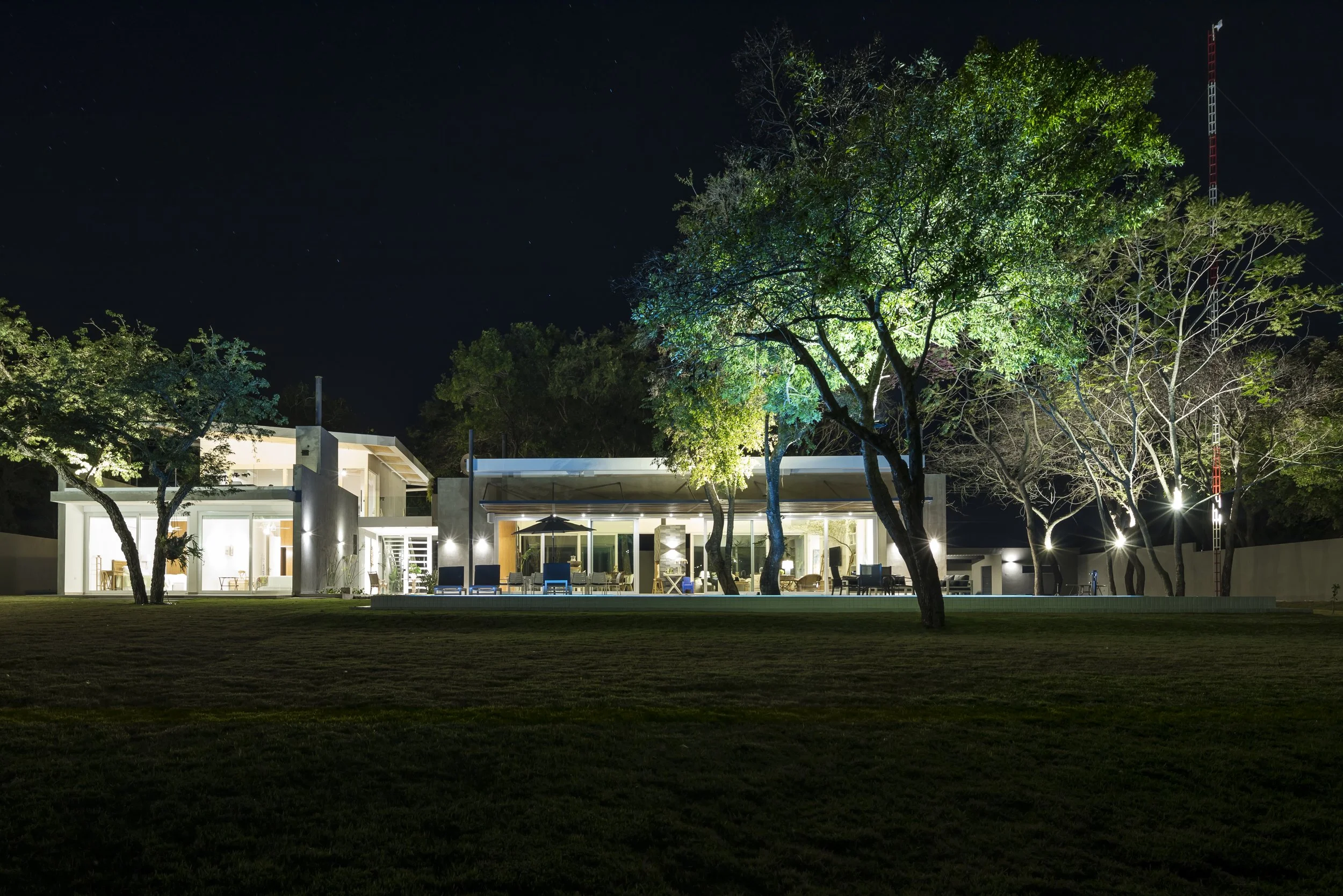 Casa moderna iluminada en la noche, rodeada de árboles, con jardín amplio y cielo estrellado.