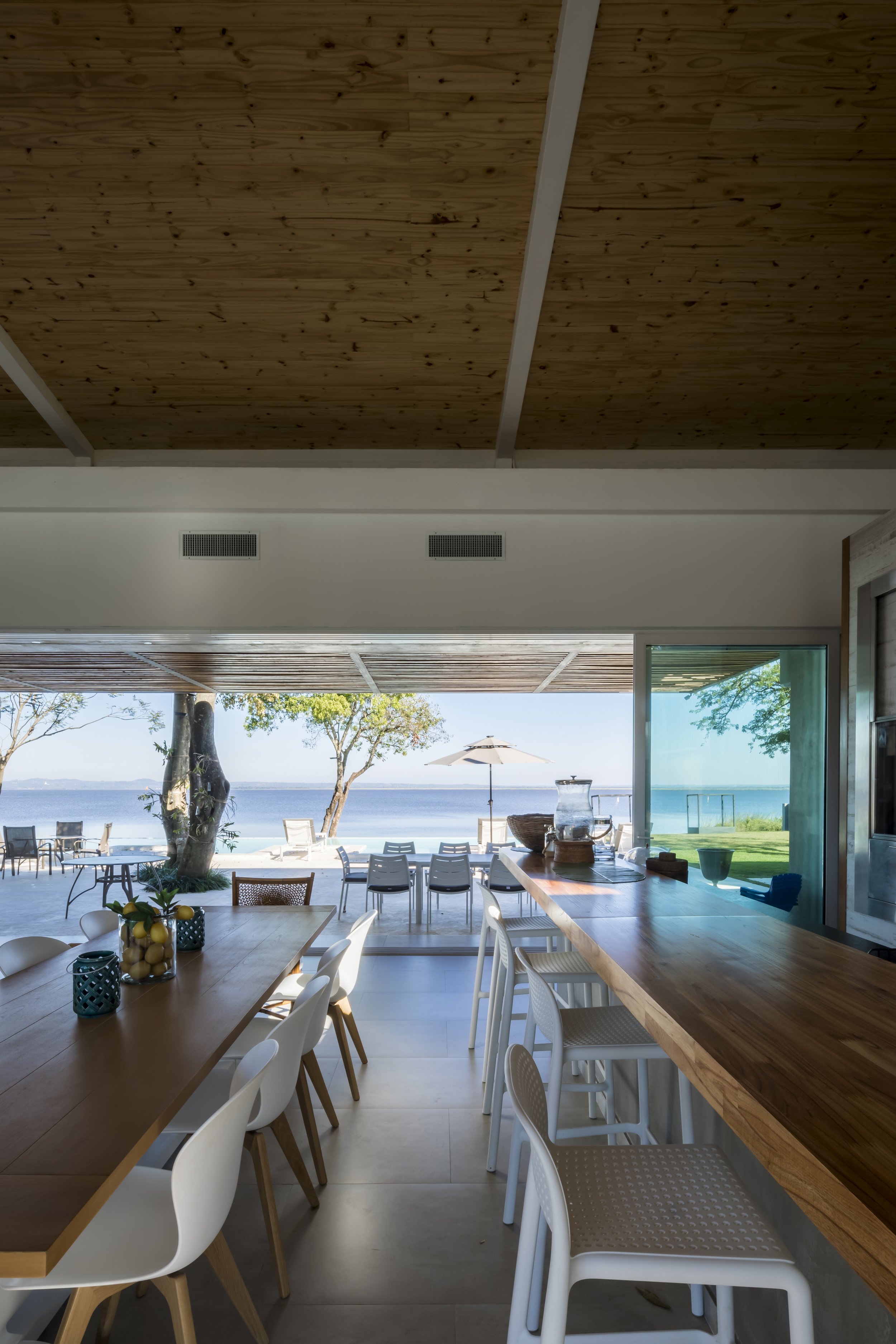 	
PRO ARQUITECTURA PARAGUAY architecture design PROSA
Vista del interior de un comedor con vista al mar, mesas y sillas blancas y de madera, bar con barra de madera, y terraza con sombrilla bajo un cielo despejado.