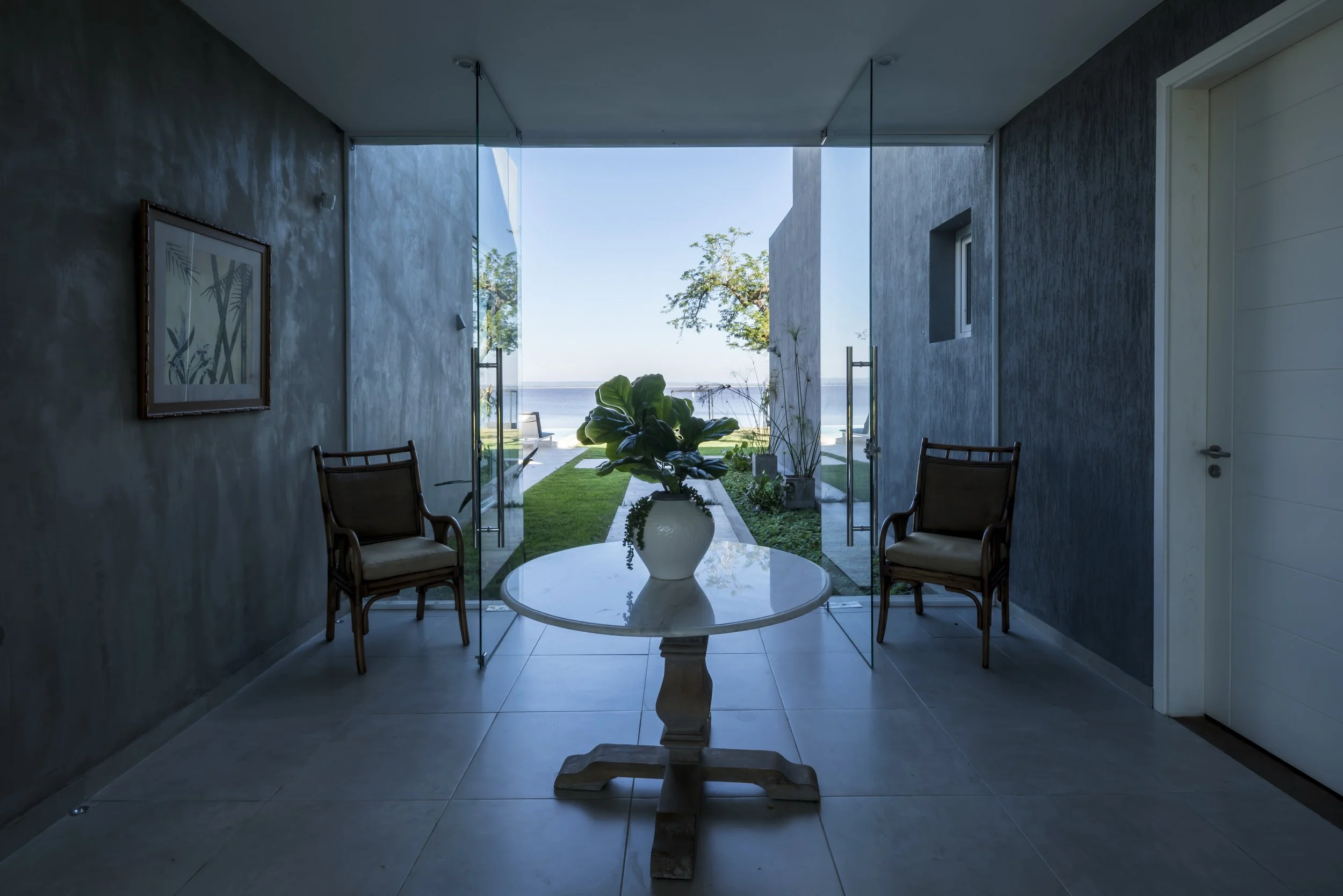 	
PRO ARQUITECTURA PARAGUAY architecture design PROSA
Vista de una sala con puertas de cristal abiertas, mostrando un jardín con vista al mar, una mesa de madera con una planta en un jarrón y dos sillas de madera.