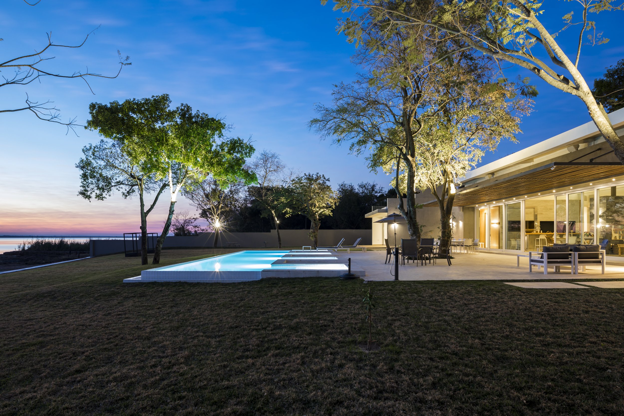 Casa moderna con piscina iluminada en la noche, rodeada de árboles y mobiliario exterior, vista al atardecer.